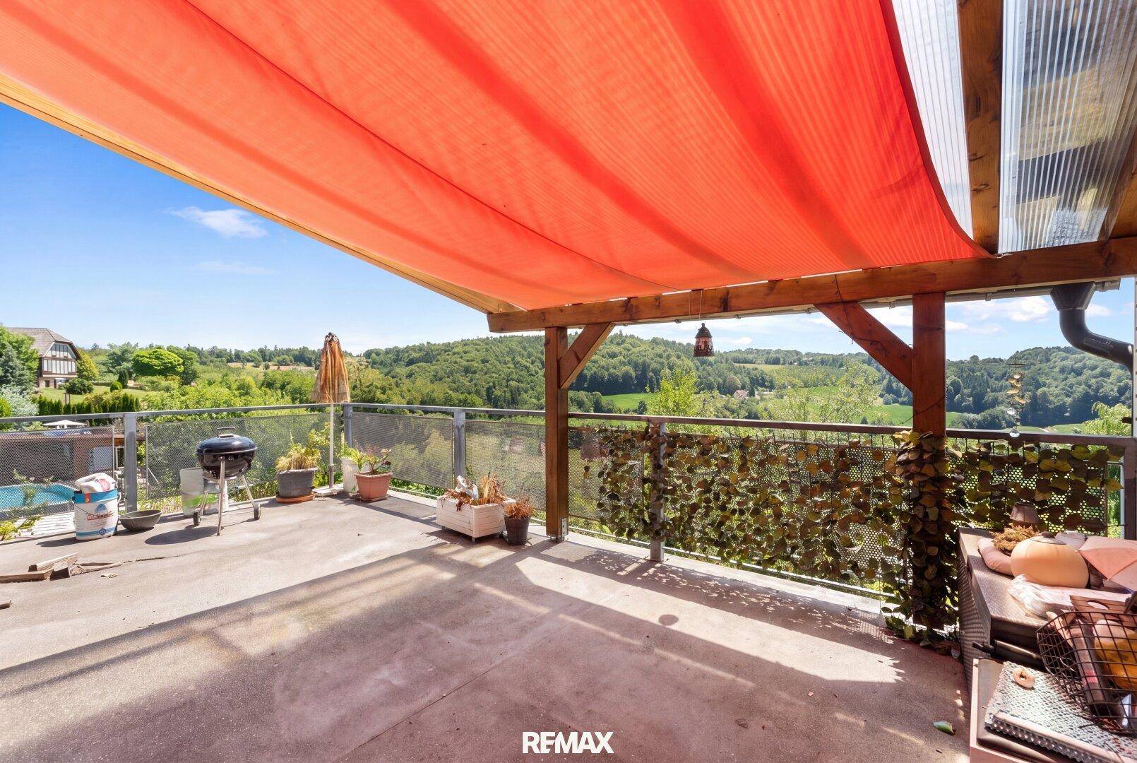Große Terrasse mit rotem Sonnensegel, Grillplatz und weitem Panoramablick über die grüne Landschaft.