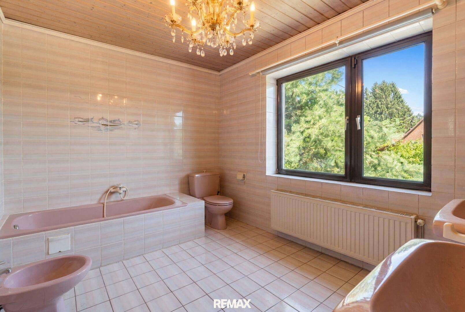 Geräumiges Badezimmer mit rosa Badewanne, Bidet und WC, umgeben von hellen Fliesen und einem Fenster.