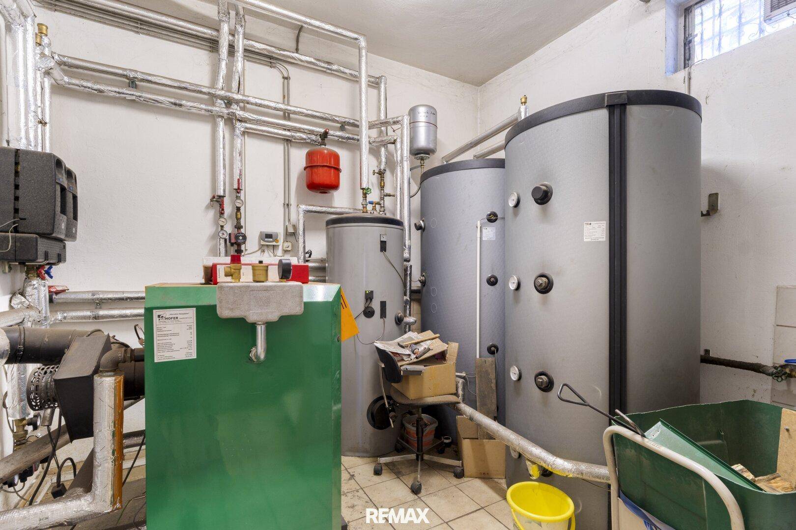 Technikraum mit großen Warmwasserspeichern, Heizungsanlage und zahlreichen Rohrleitungen auf Fliesenboden.