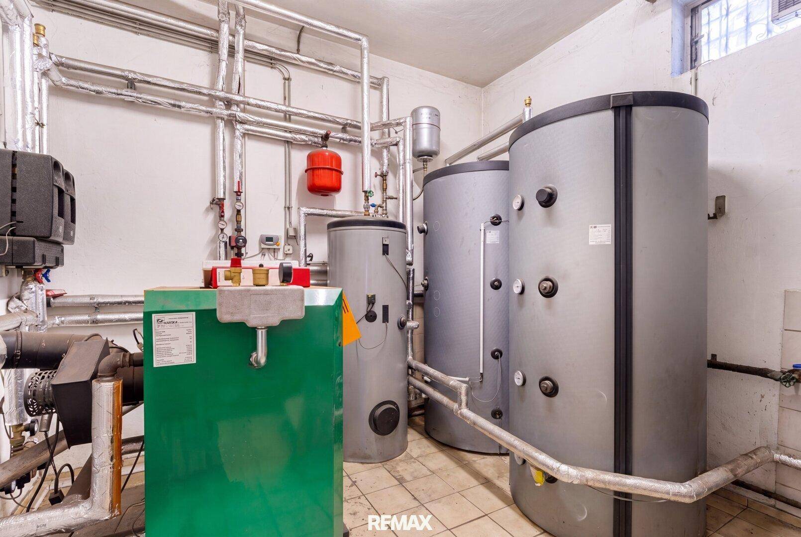 Technikraum mit Heizungsanlage, Warmwasserspeichern und komplexer Rohrinstallation auf Fliesenboden.