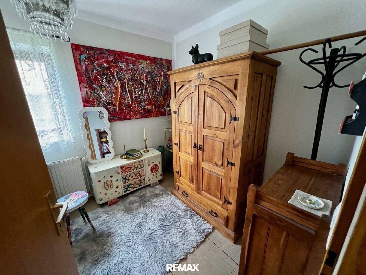 Kleines Schlafzimmer mit Holzboden, traditionellem Holzschrank und farbenfroher Wandkunst.