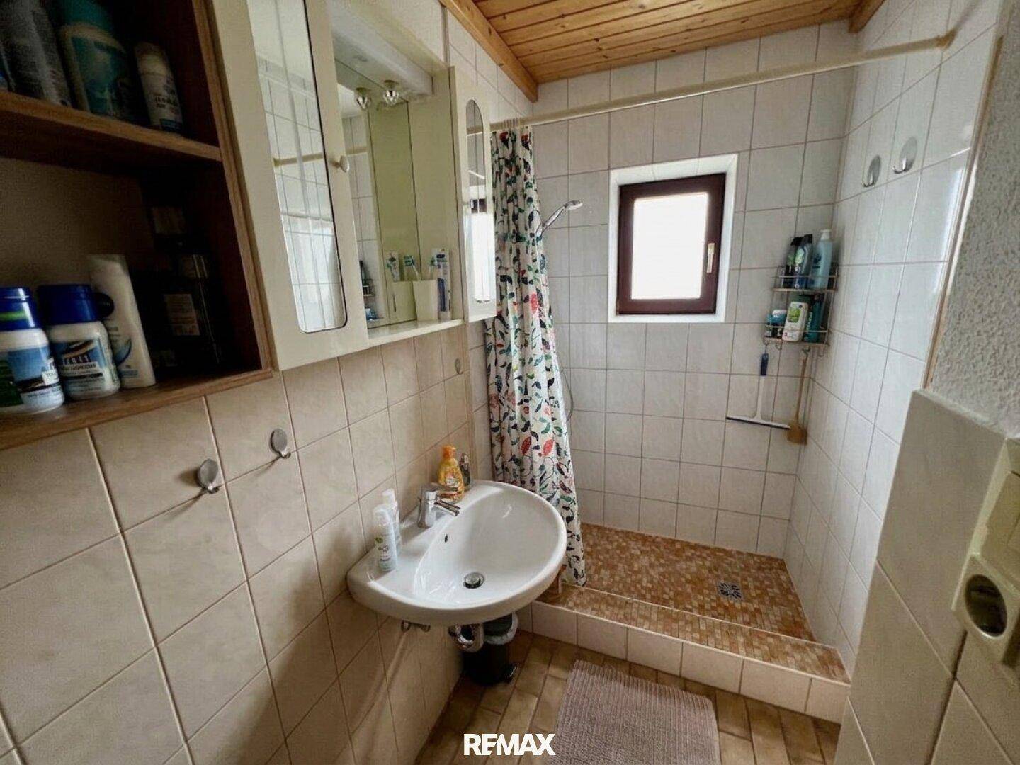 Einfaches Badezimmer mit gefliesten Wänden, Duschbereich, Waschbecken und Holzdecke.