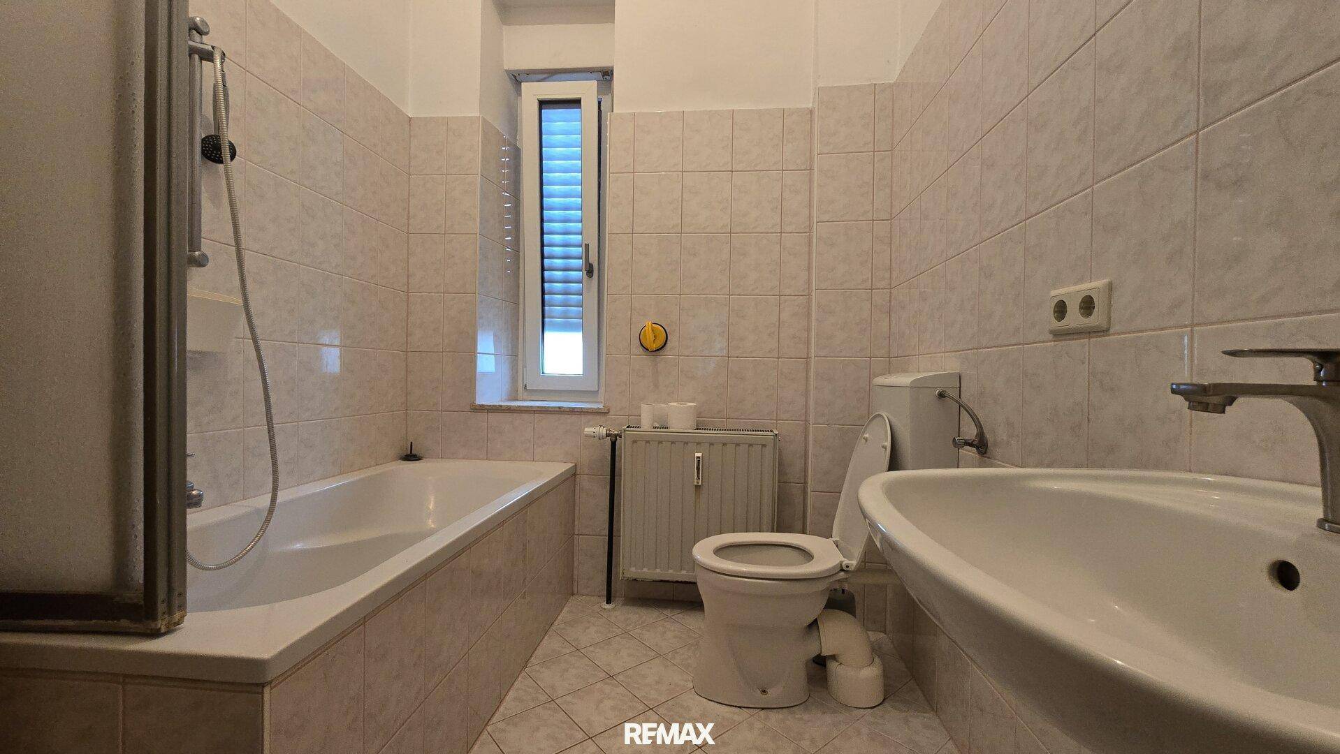 Gefliestes Badezimmer mit Badewanne, Toilette, Waschbecken und Fenster für natürliche Belüftung.
