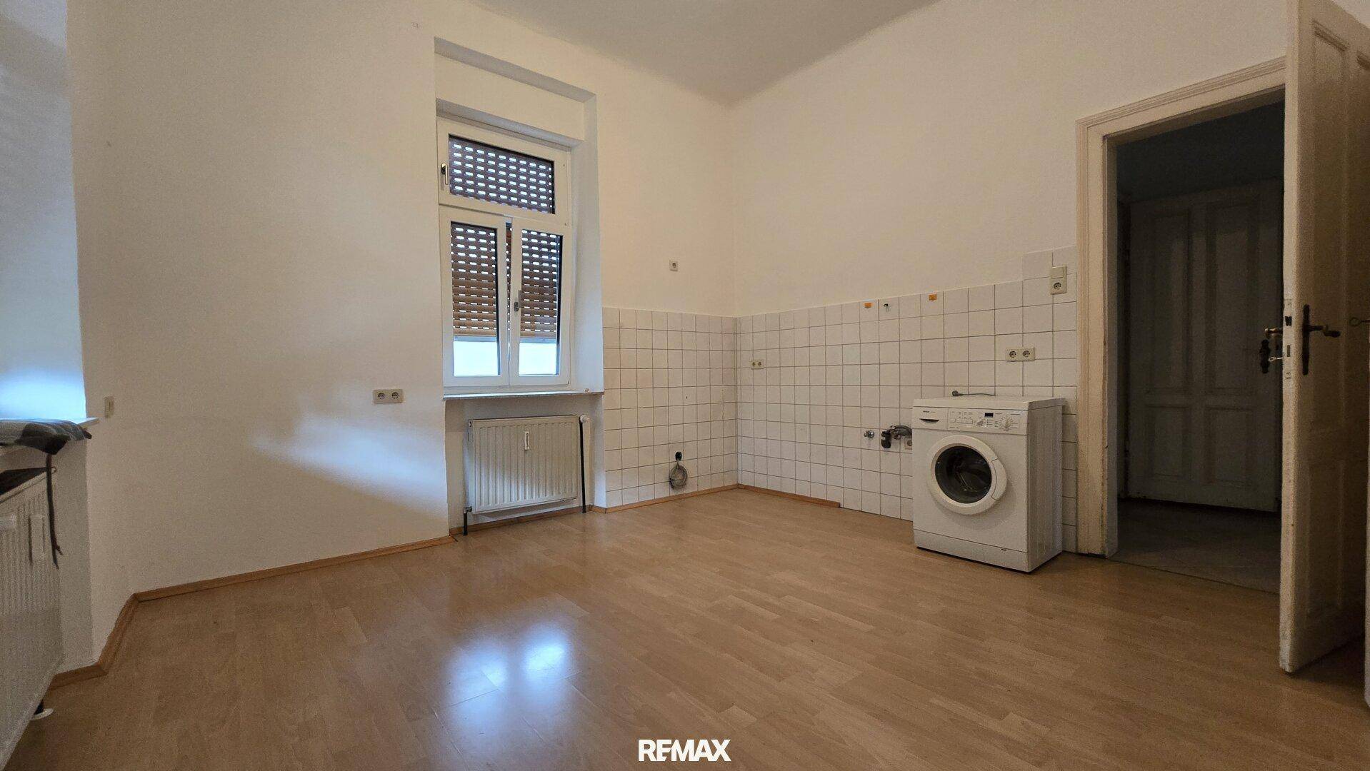 Leere Küche mit Fliesenwand, Waschmaschinenanschluss und Fenster mit Rollladen.
