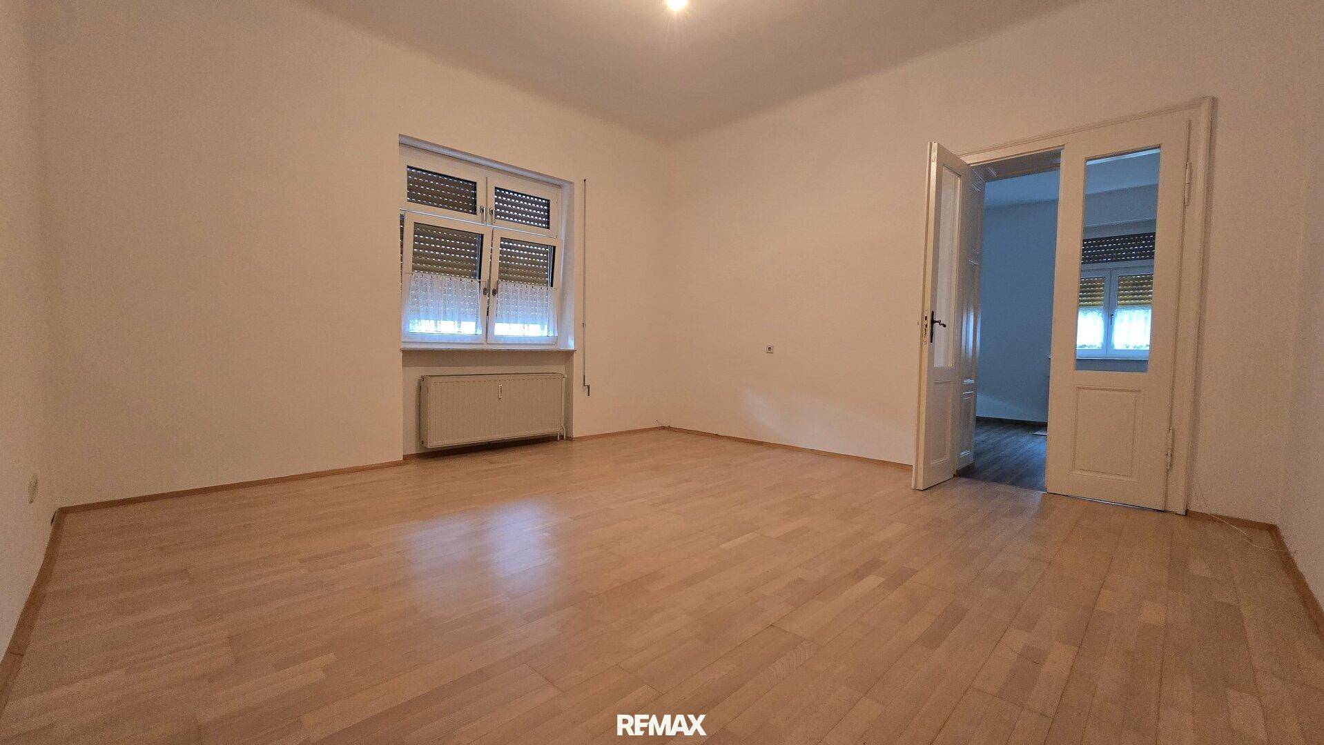 Geräumiges Wohnzimmer mit hellem Laminatboden und Doppeltüren zu einem weiteren Raum.