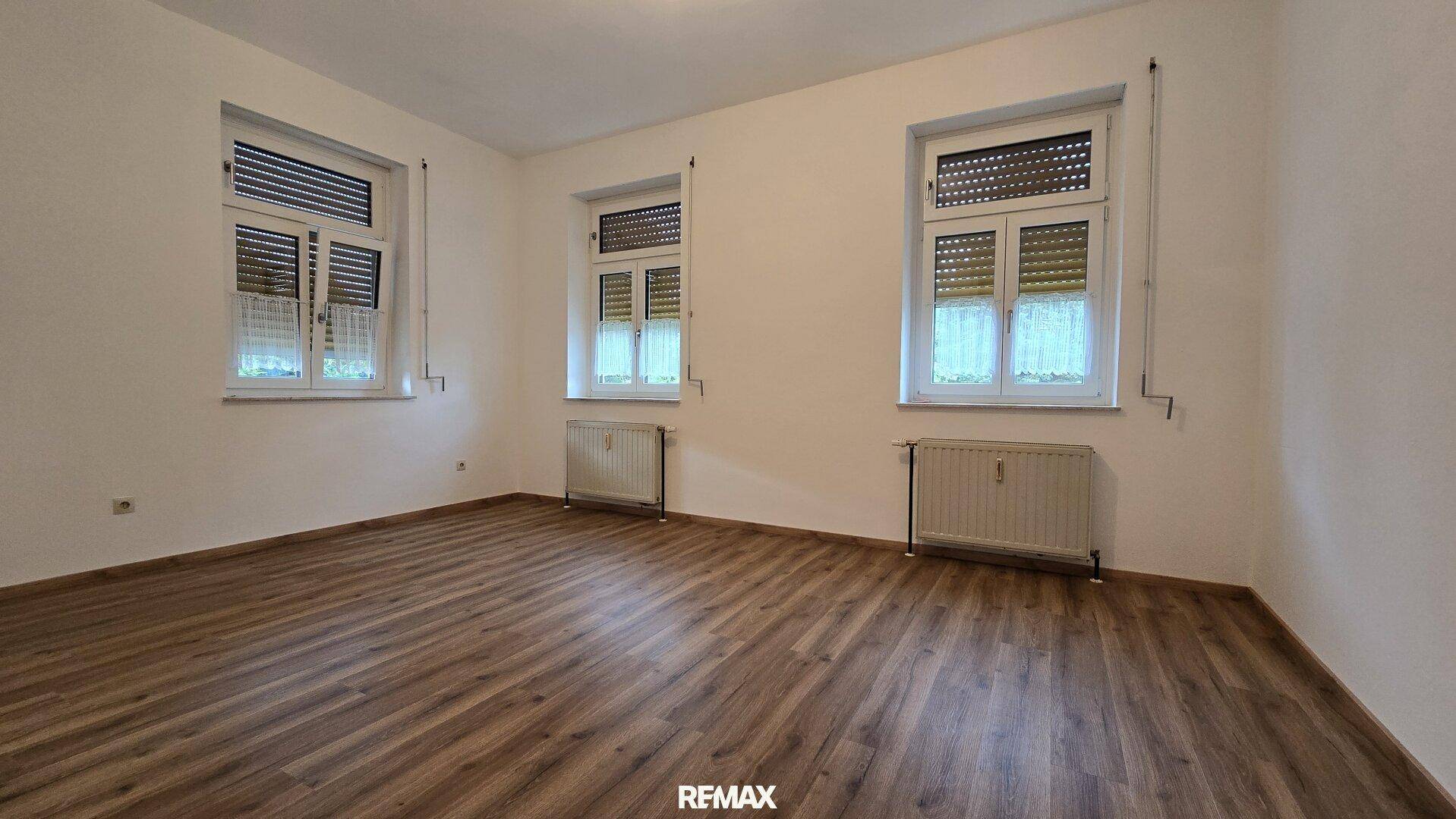 Helles Wohnzimmer mit Laminatboden und drei Fenstern, die viel Tageslicht hereinlassen.