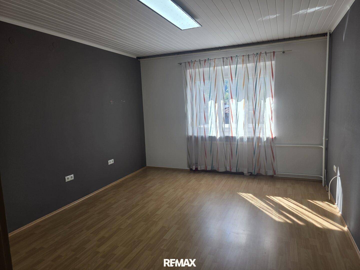 Leeres Zimmer mit Holzboden, dunklen Wänden und einem Fenster mit bunten Vorhängen.