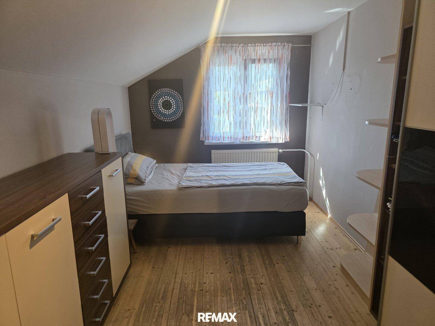 Gemütliches Schlafzimmer mit Holzboden, Dachschräge und einem Fenster mit Vorhängen.