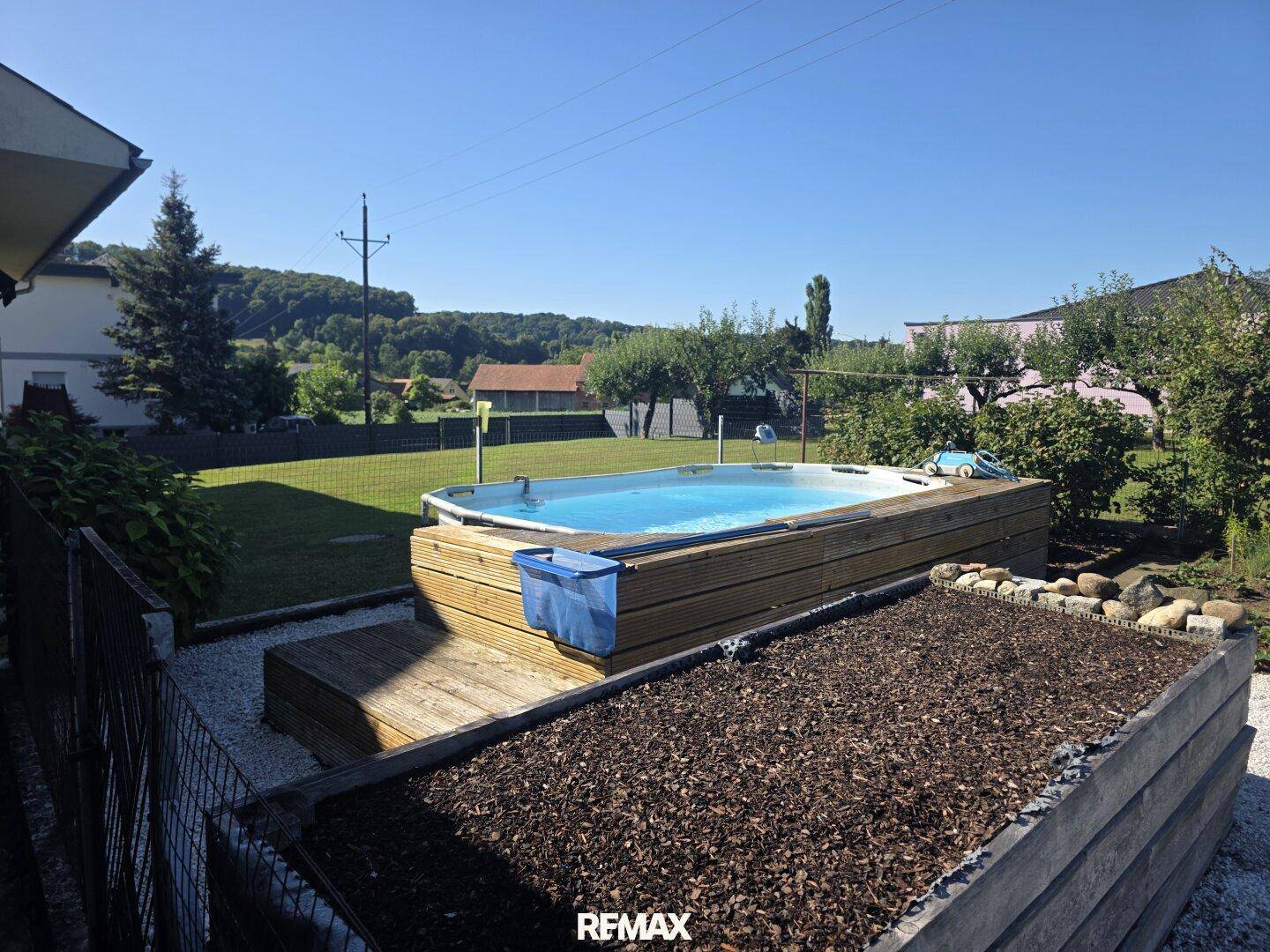 Gartenansicht mit dem Poolbereich und einem Hochbeet, umgeben von Rasen und Natur.