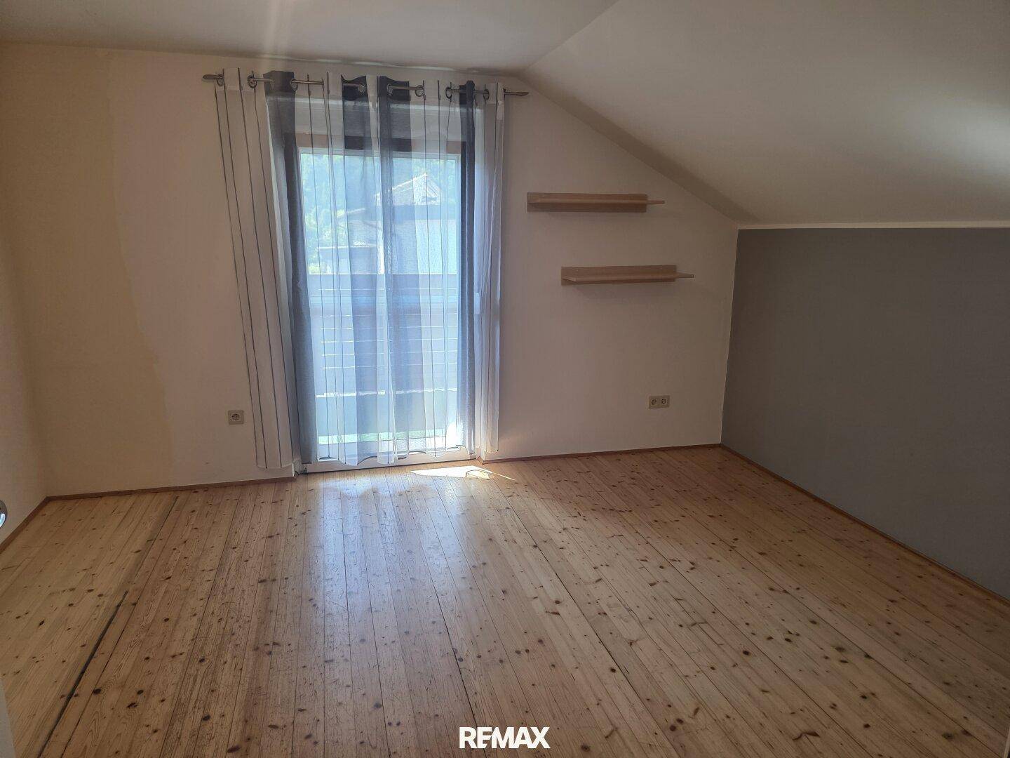 Helles Zimmer mit Holzboden, Dachschräge und Zugang zu einem Balkon.