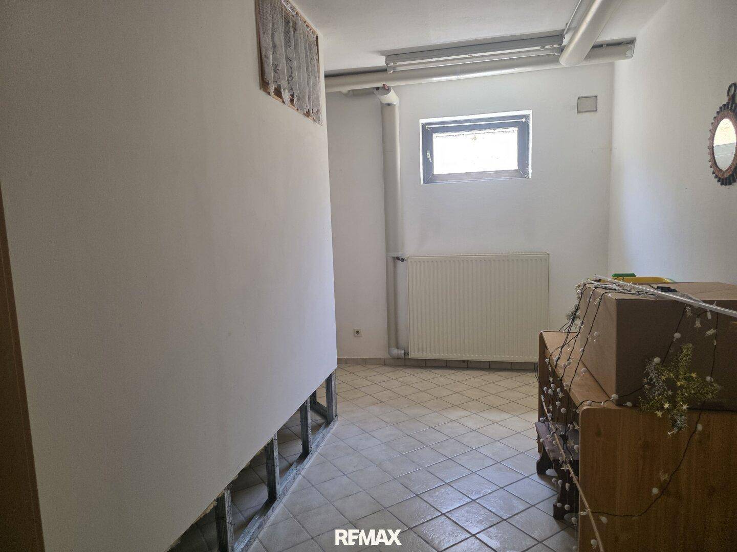 Unfertiger Kellerraum mit Fliesenboden, kleinem Fenster und Heizkörper, ideal als Abstellraum.