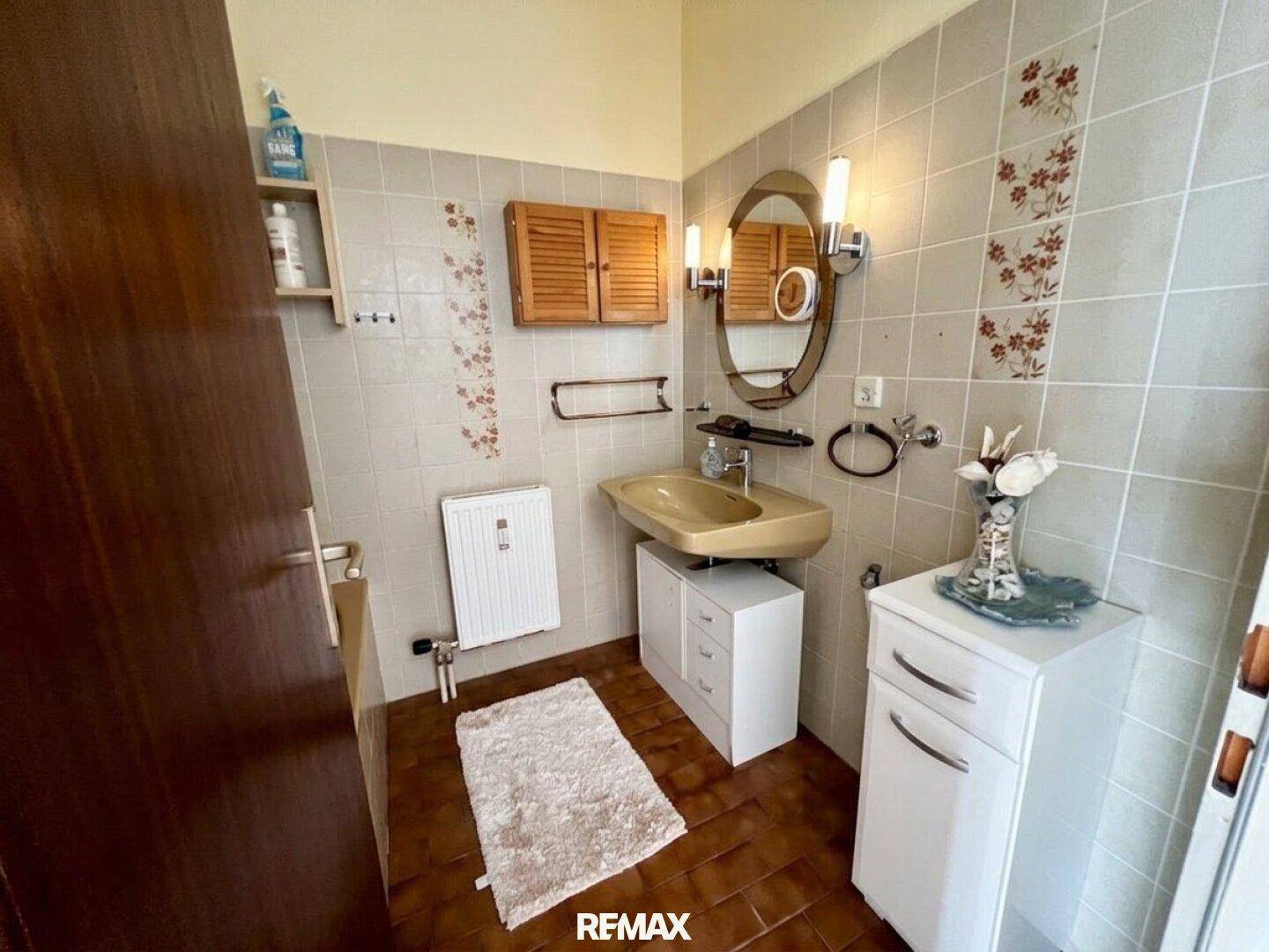 Badezimmer mit beigen Fliesen, Waschbecken, ovalem Spiegel und Holzschrank im klassischen Stil.