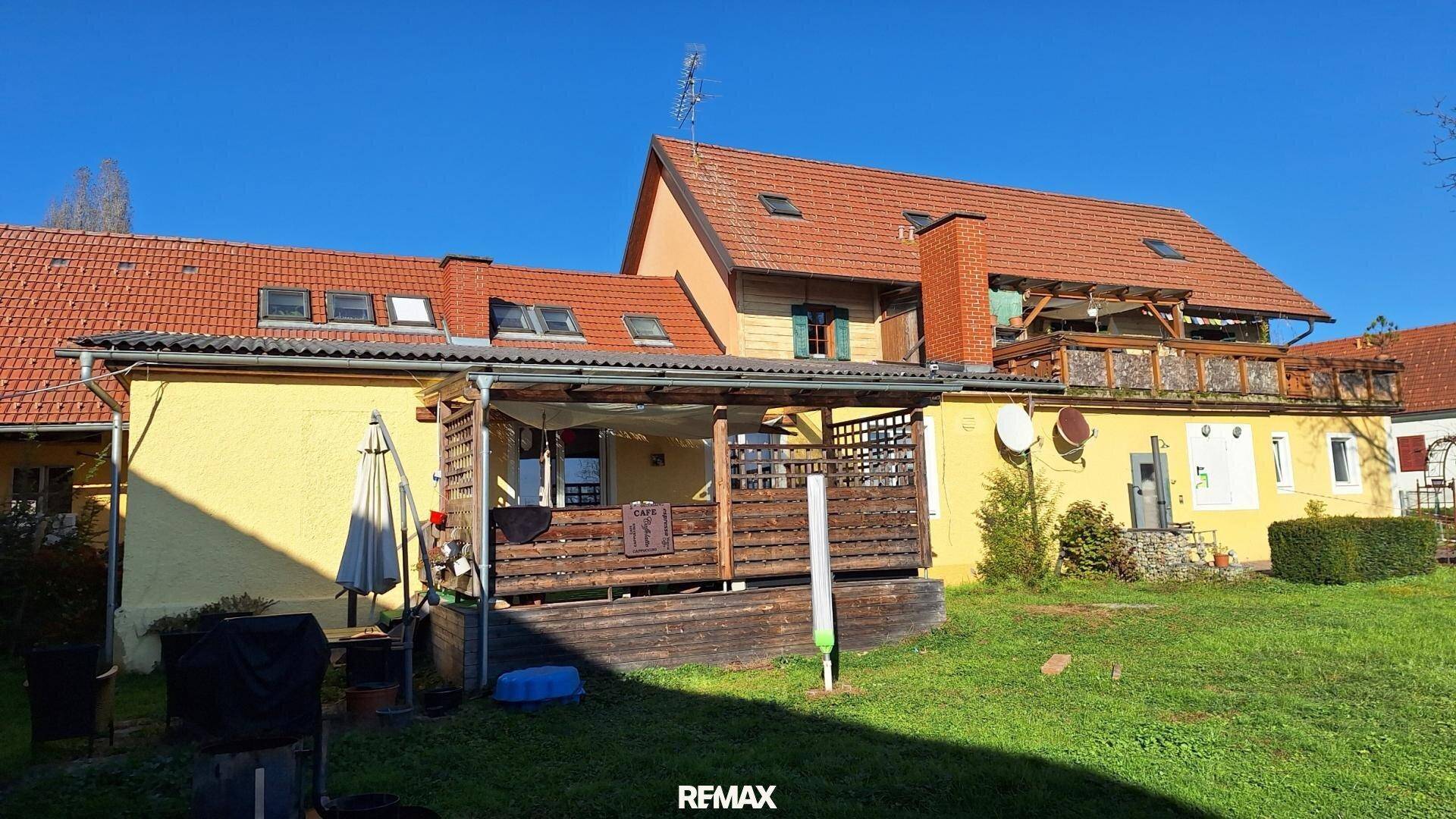 Rückseite des Hauses mit überdachter Terrasse, Garten und Dachfenstern unter blauem Himmel.