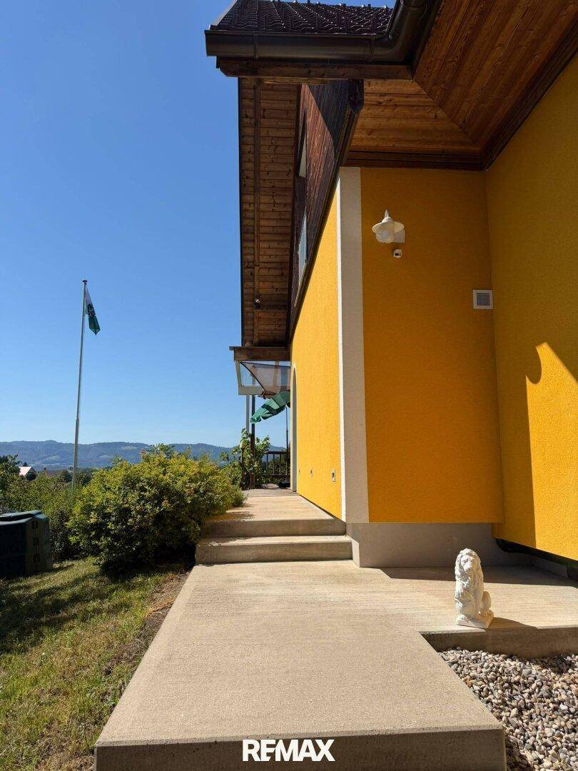Seitenansicht des Hauses mit gelber Fassade, Betonweg und Blick auf die grüne Umgebung.