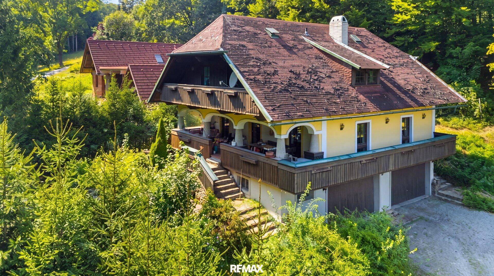 Traditionelles Haus mit gelber Fassade, Holzbalkonen und einem roten Ziegeldach, umgeben von Bäumen.