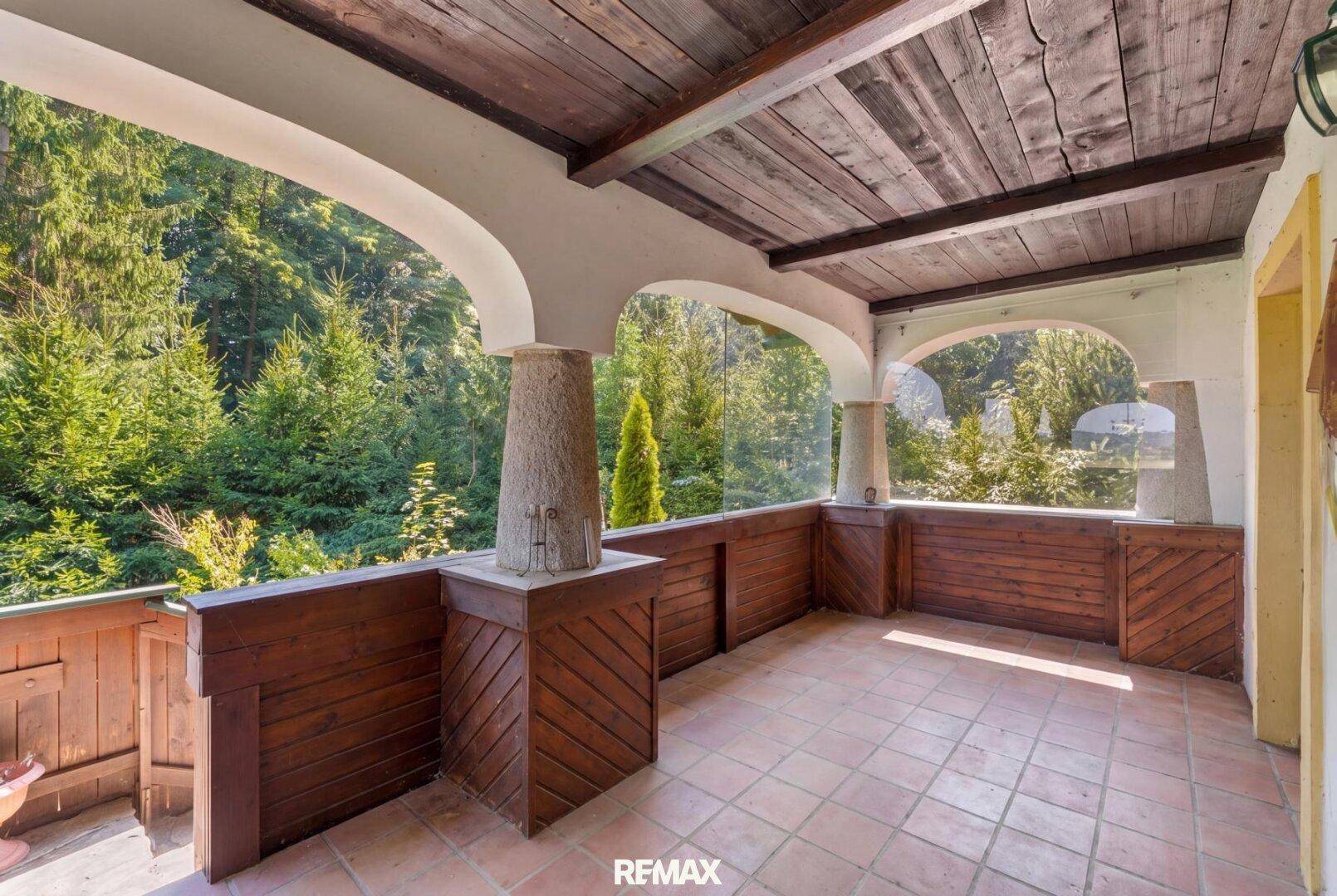 Helle Loggia mit Terrakottaboden und Holzbalkendecke, umgeben von üppigem Grün.