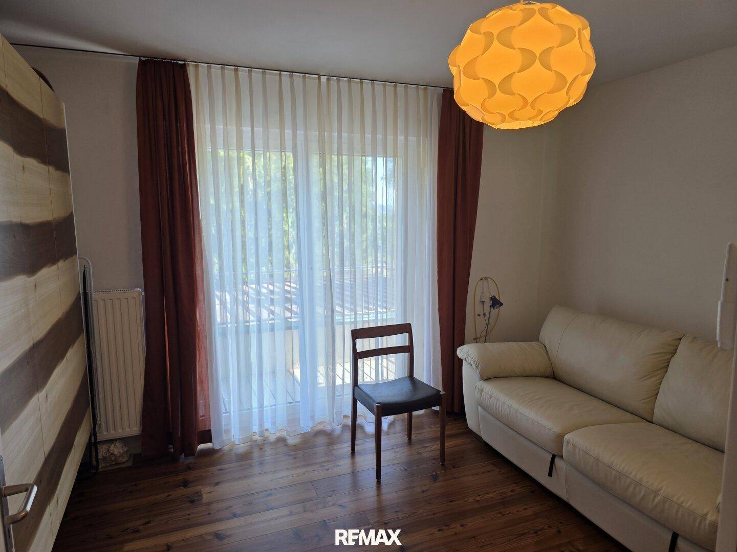 Helles Zimmer mit Holzboden, Sofa und Zugang zu einem Außenbereich durch große Fenster.