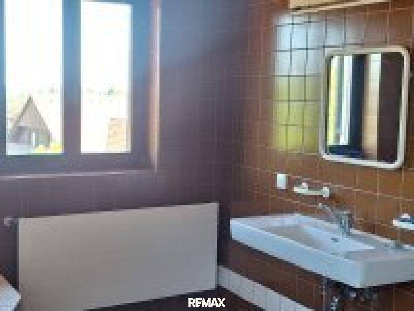 Badezimmer mit braunen Fliesen, Waschbecken, Spiegel und einem großen Fenster für Tageslicht.
