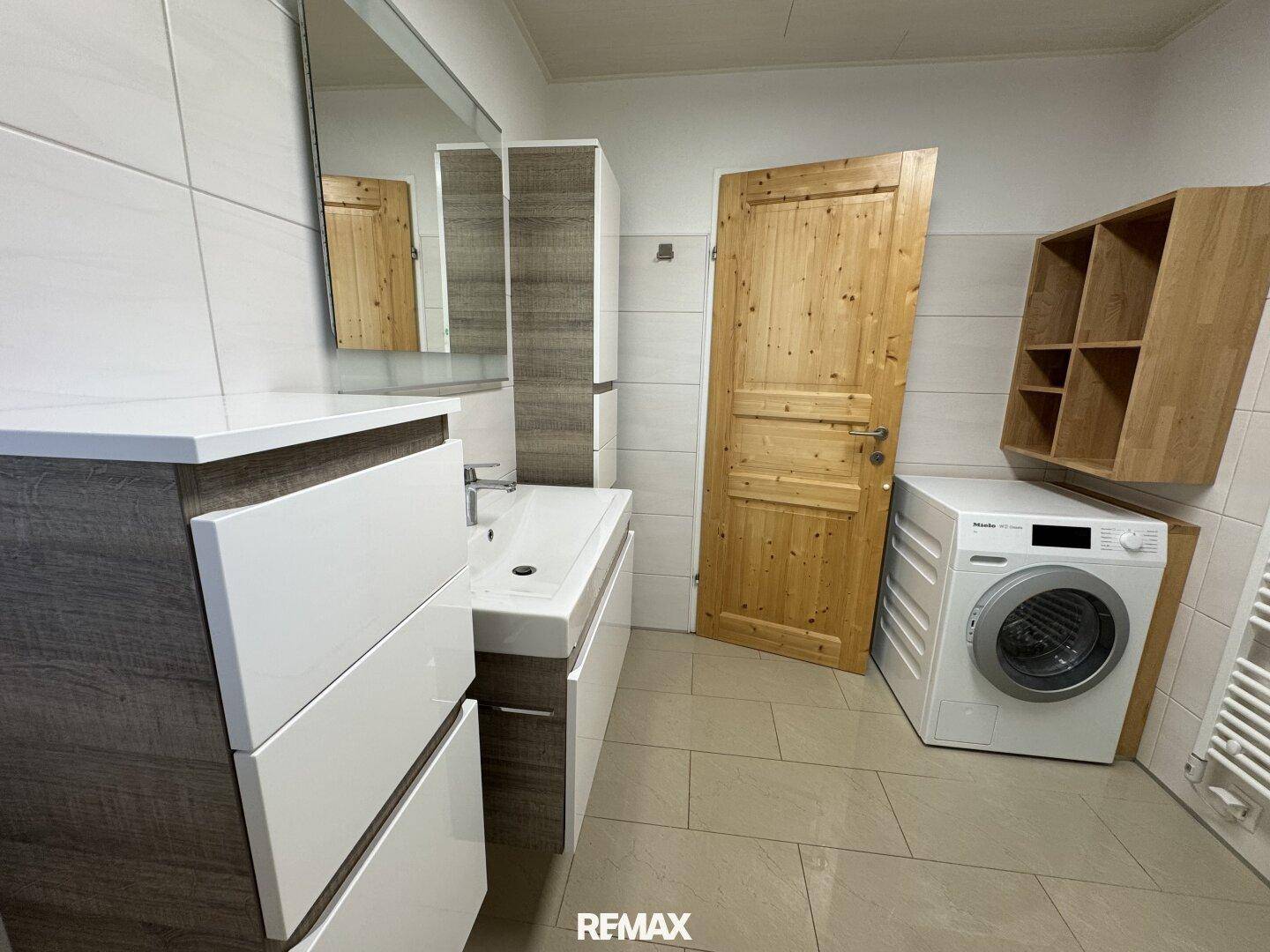 Badezimmer mit Waschmaschine, Waschtisch und Holzregal, hell gefliest.