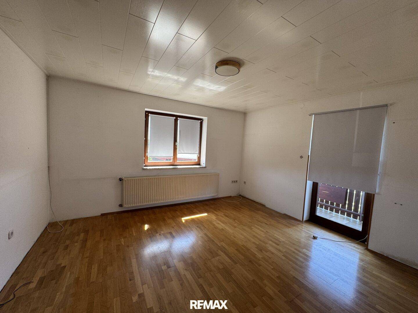 Geräumiges, leeres Zimmer mit Holzboden, großem Fenster und Zugang zum Balkon.