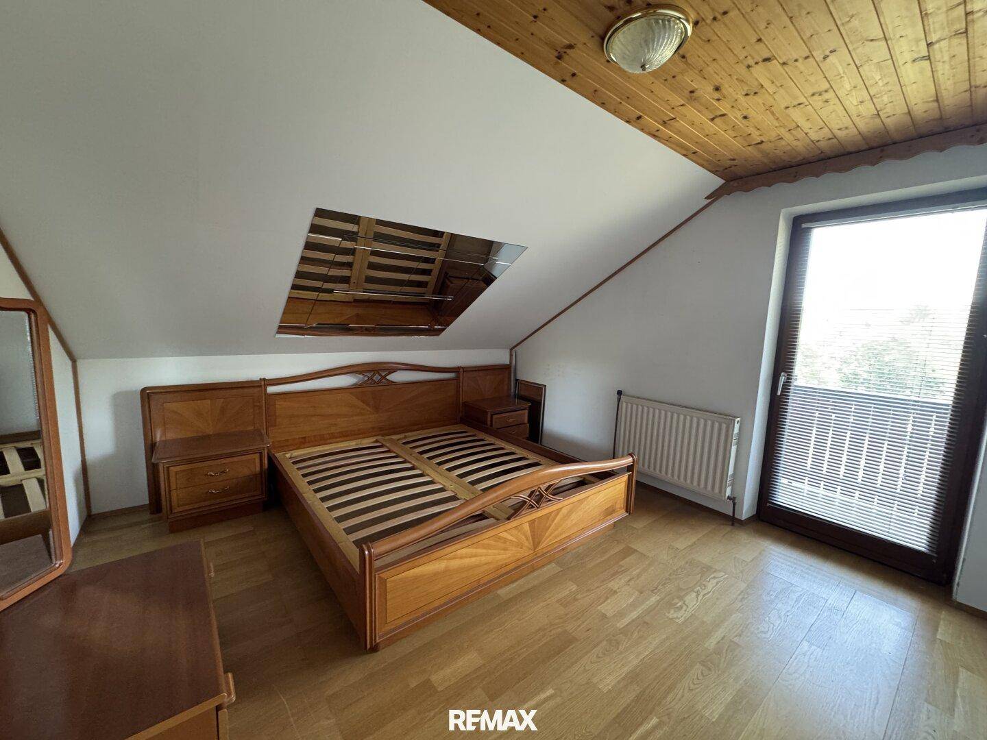 Gemütliches Schlafzimmer mit Holzmöbeln, Holzdecke, Schrägdach und großem Fenster mit Blick nach draußen.