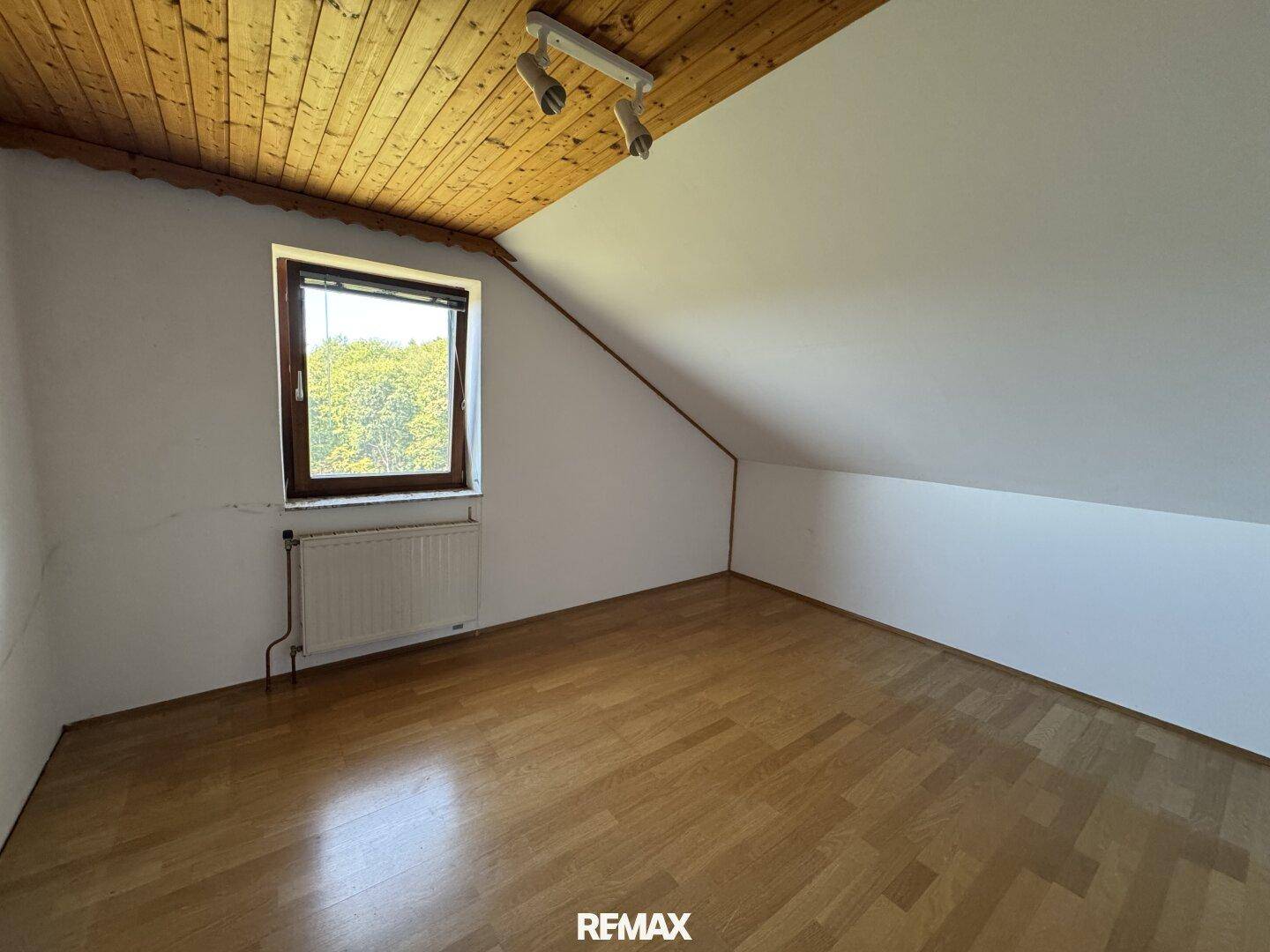 Helles Dachzimmer mit Holzdecke, Schrägdach und Fenster, ausgestattet mit Heizkörper und Holzfußboden.