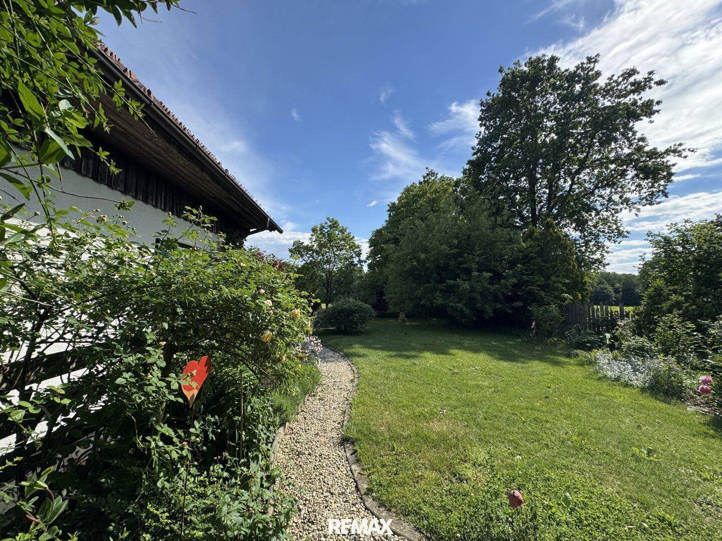 Weitläufiger Garten mit Rasenfläche, Kiesweg und altem Baumbestand unter blauem Himmel.