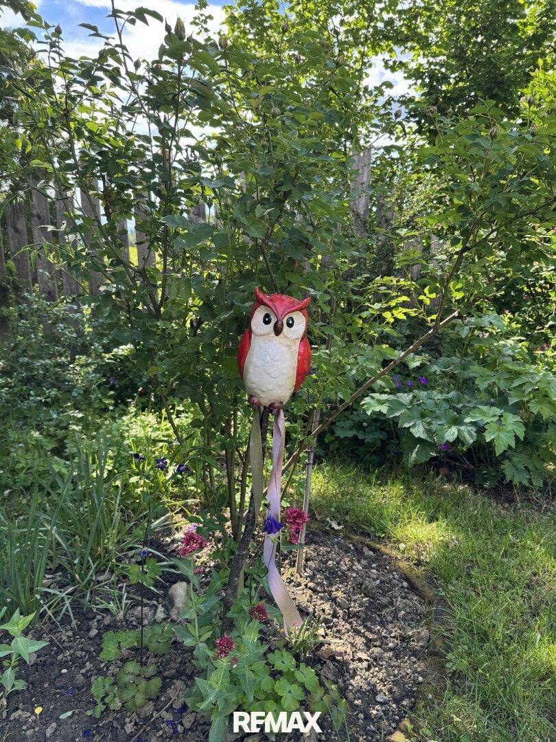Dekorative Eule auf einem Pfahl in einem üppigen Garten mit verschiedenen Pflanzen und Bäumen.