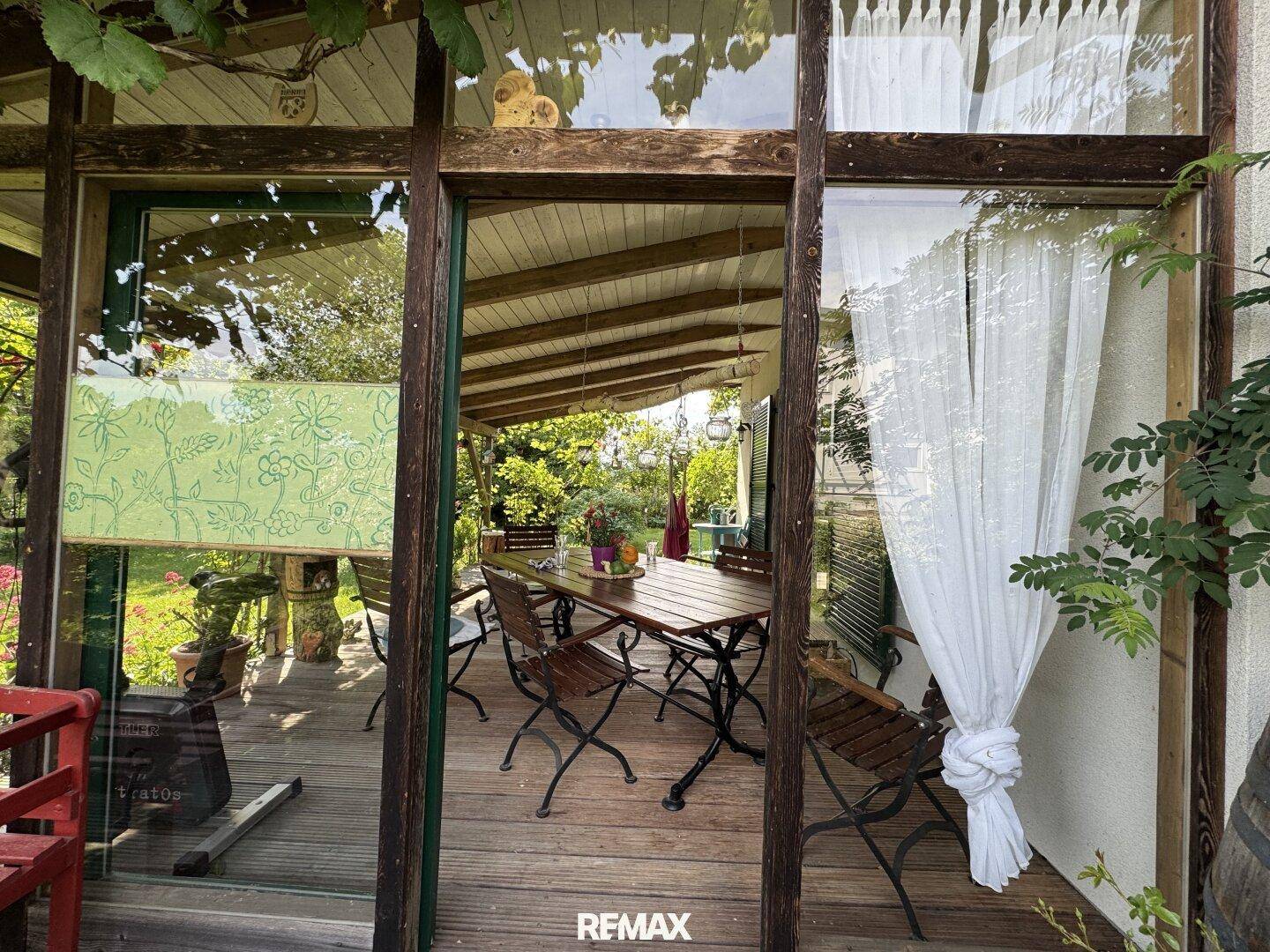 Blick vom Innenraum durch große Fenster auf die überdachte Holzterrasse mit Esstisch.