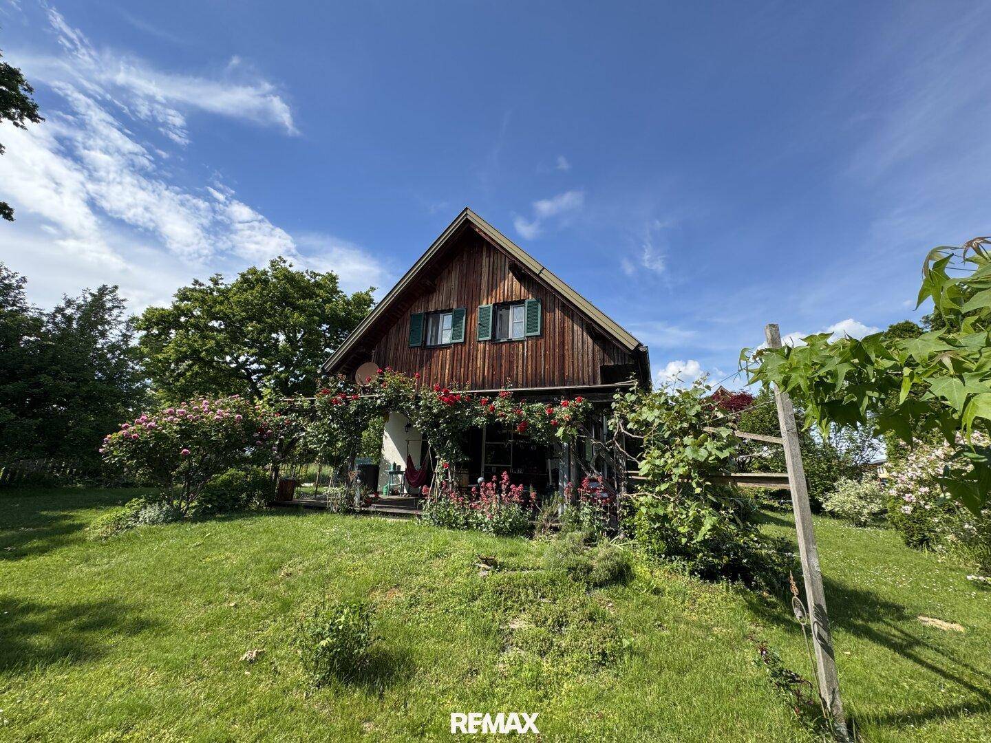 Weitläufiger Blick auf das charmante Holzhaus mit blühenden Rosen und großem Garten unter blauem Himmel.