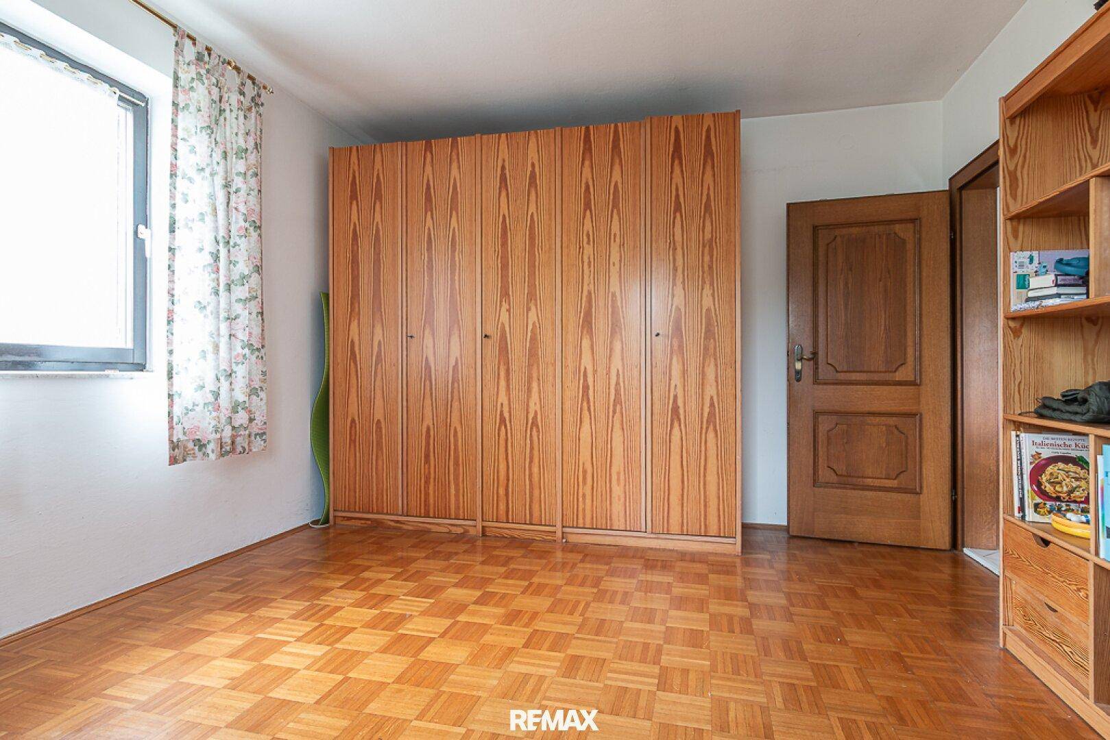 Schlafzimmer mit Parkettboden, großem Holzschrank und Fenster mit floralen Vorhängen.