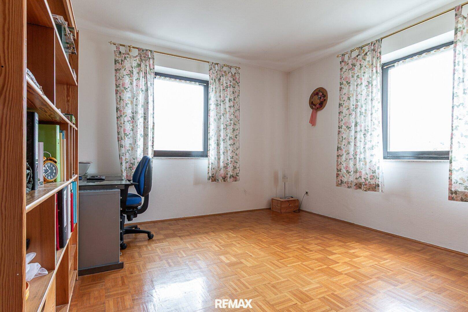 Helles Arbeitszimmer mit Parkettboden, Bücherregal und zwei Fenstern.