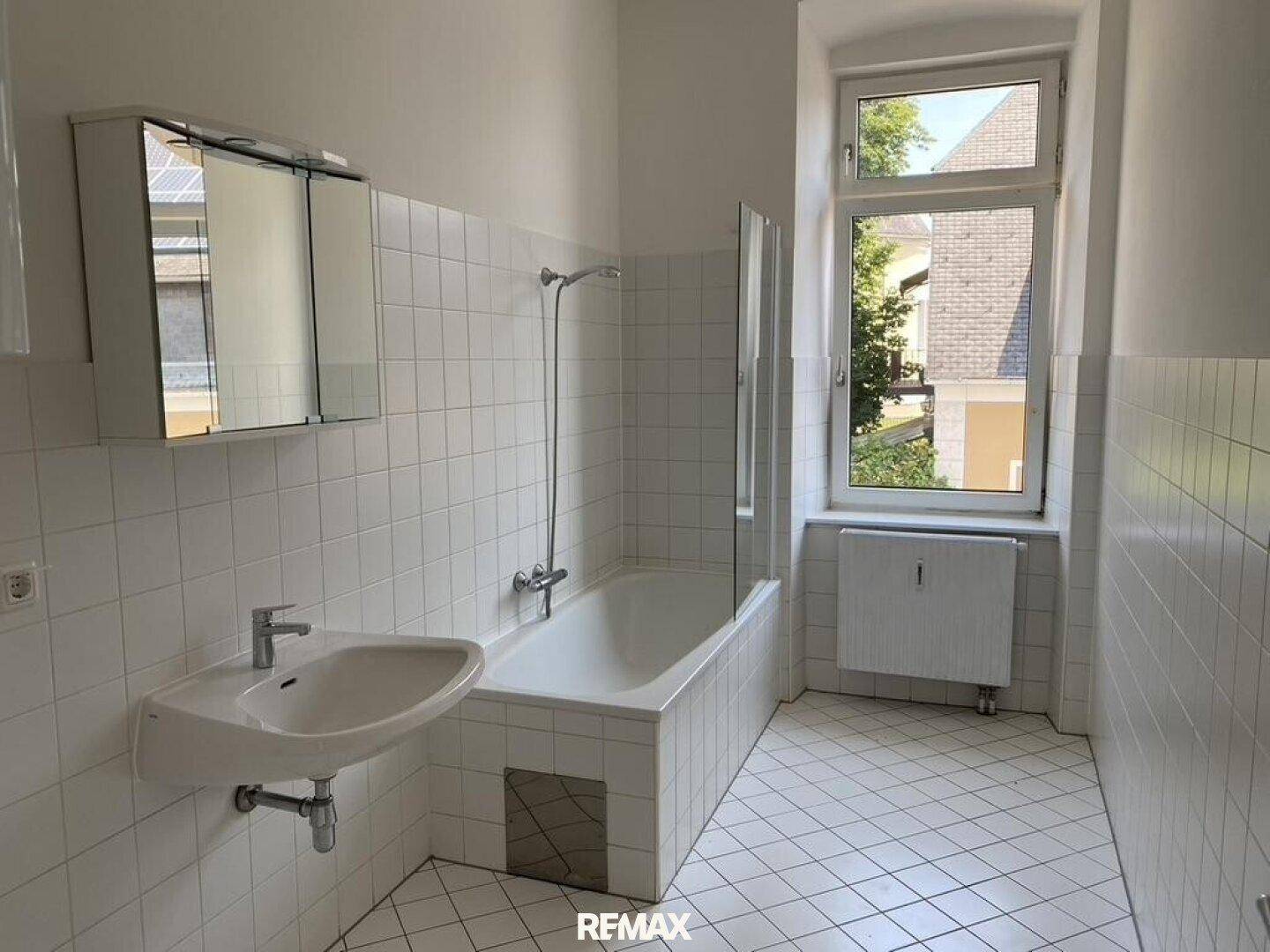Badezimmer mit weißen Fliesen, Badewanne mit Duschfunktion, Waschbecken und Spiegelschrank.