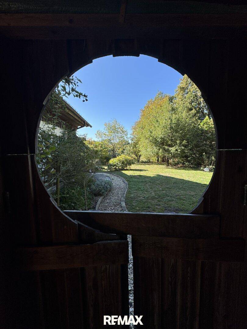 Malerischer Blick durch eine runde Öffnung in einem Holzschuppen auf den sonnigen Gartenweg.