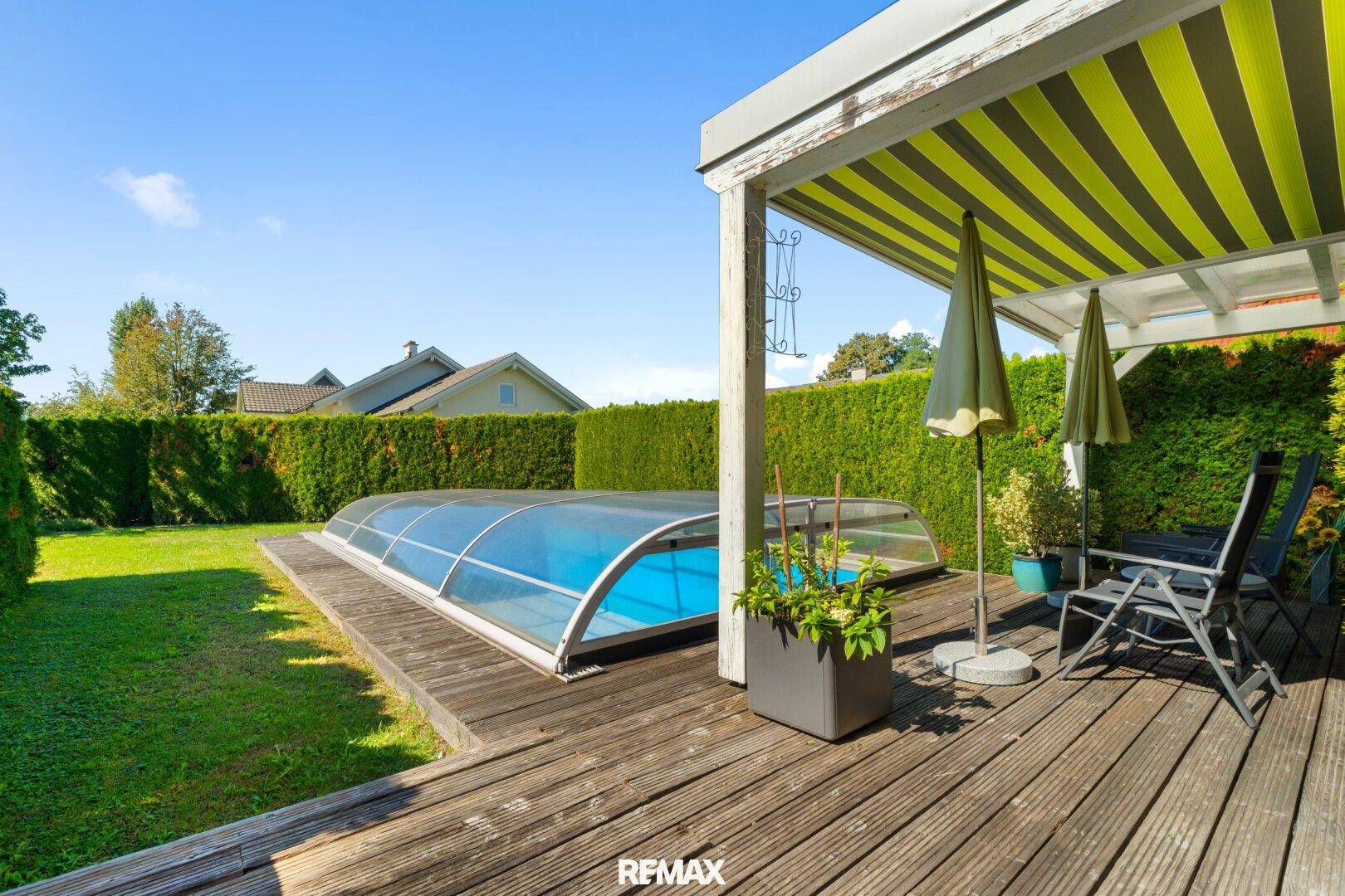 Holzterrasse mit überdachtem Pool, Gartenmöbeln und umgebender grüner Hecke.
