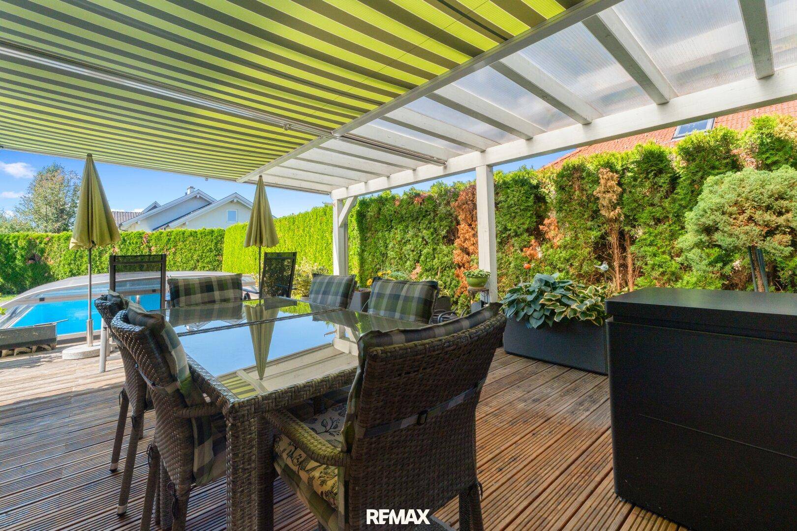 Überdachte Holzterrasse mit großem Esstisch und Blick auf den Pool und den Garten.