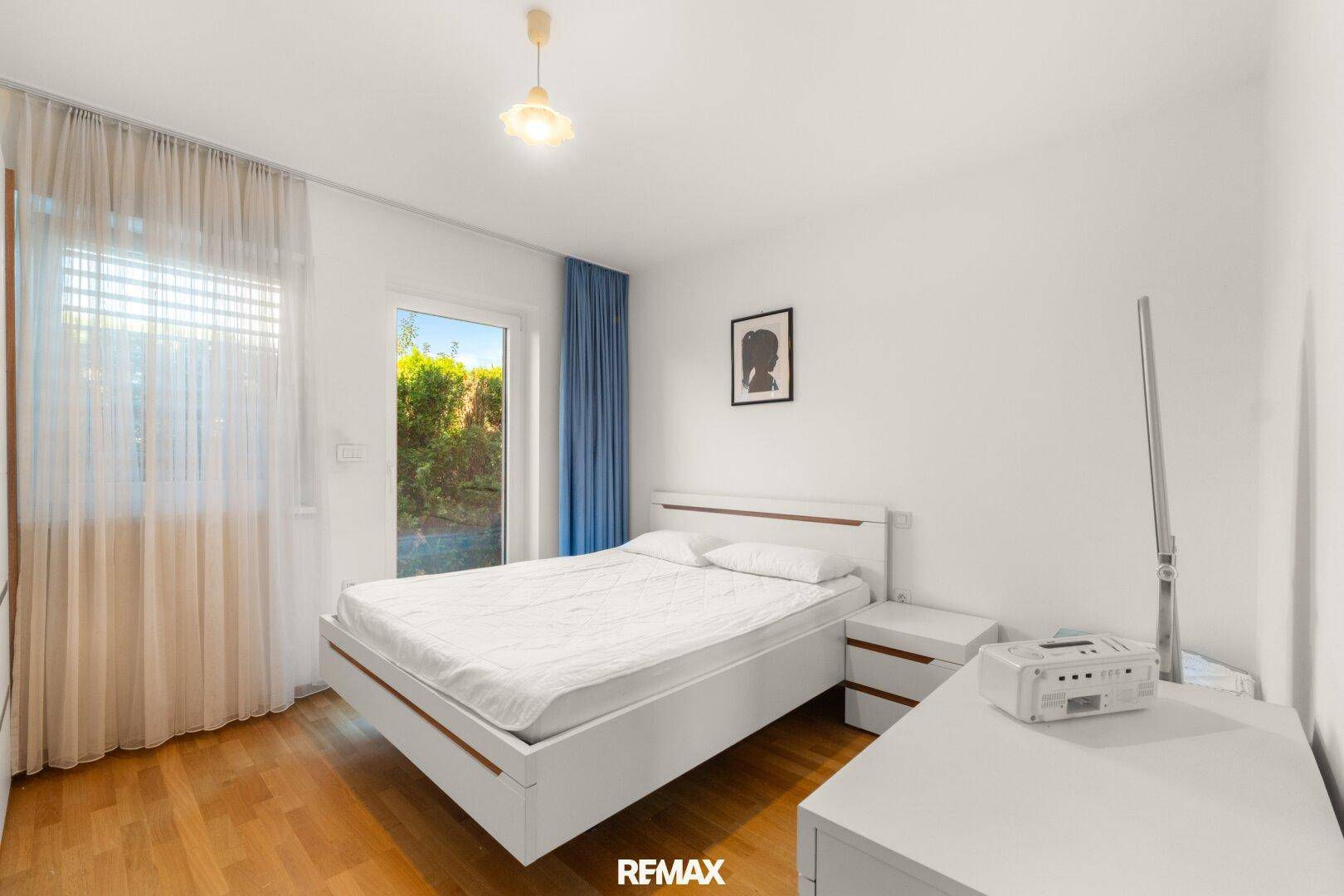 Schlafzimmer mit Holzboden, weißem Bett und Fenster mit Blick ins Grüne.