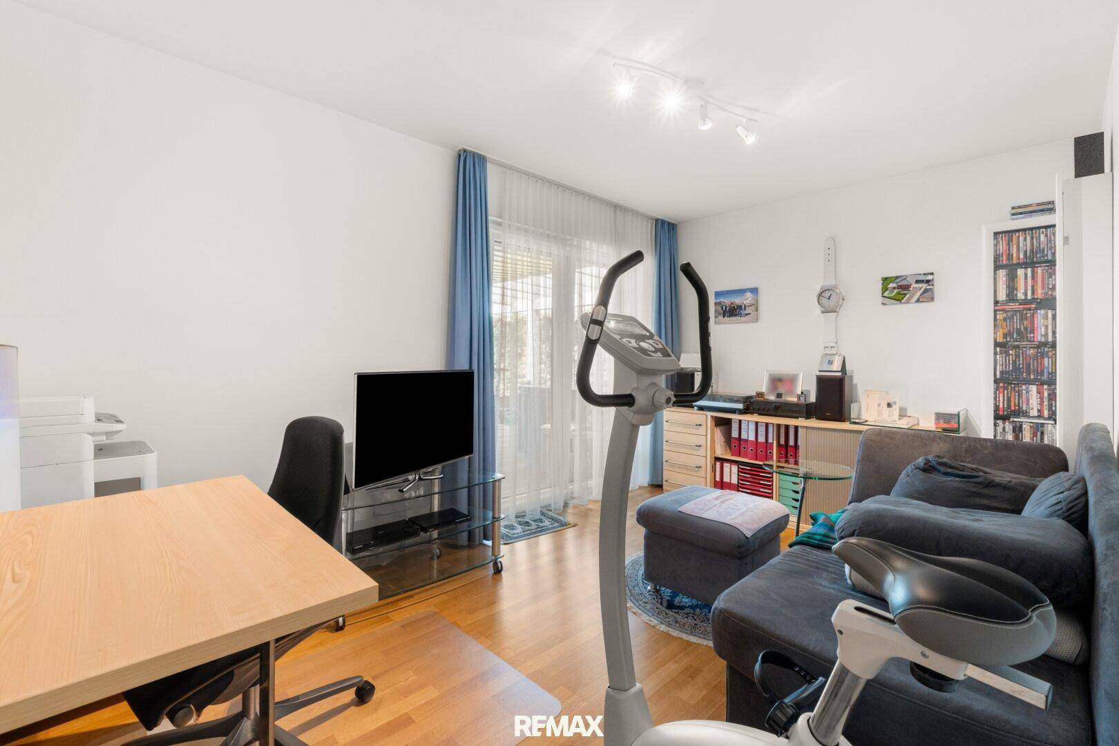 Vielseitiger Wohnraum mit Sofa, Schreibtisch und Fitnessgerät, hell und funktional eingerichtet.