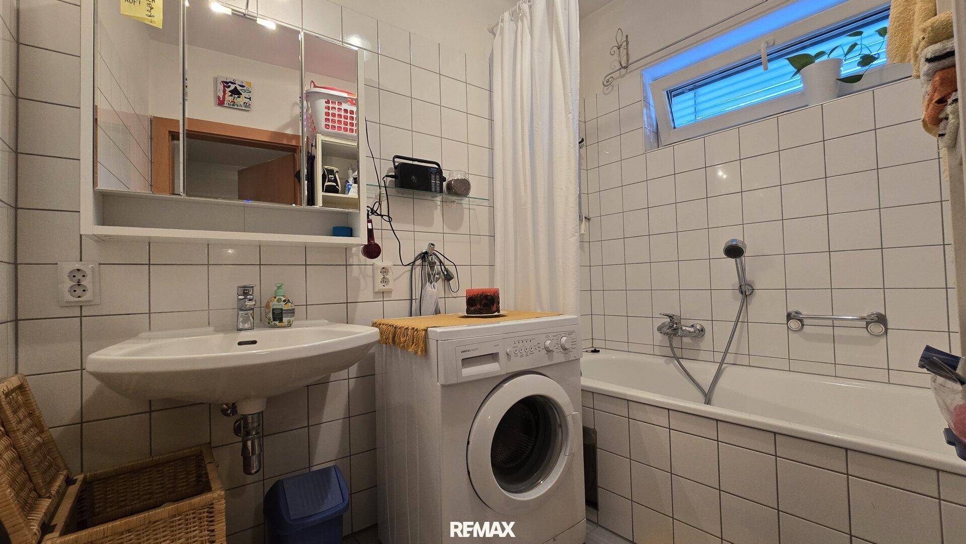 Helles Badezimmer mit weißen Fliesen, Badewanne mit Duschfunktion, Waschbecken und Spiegelschrank.