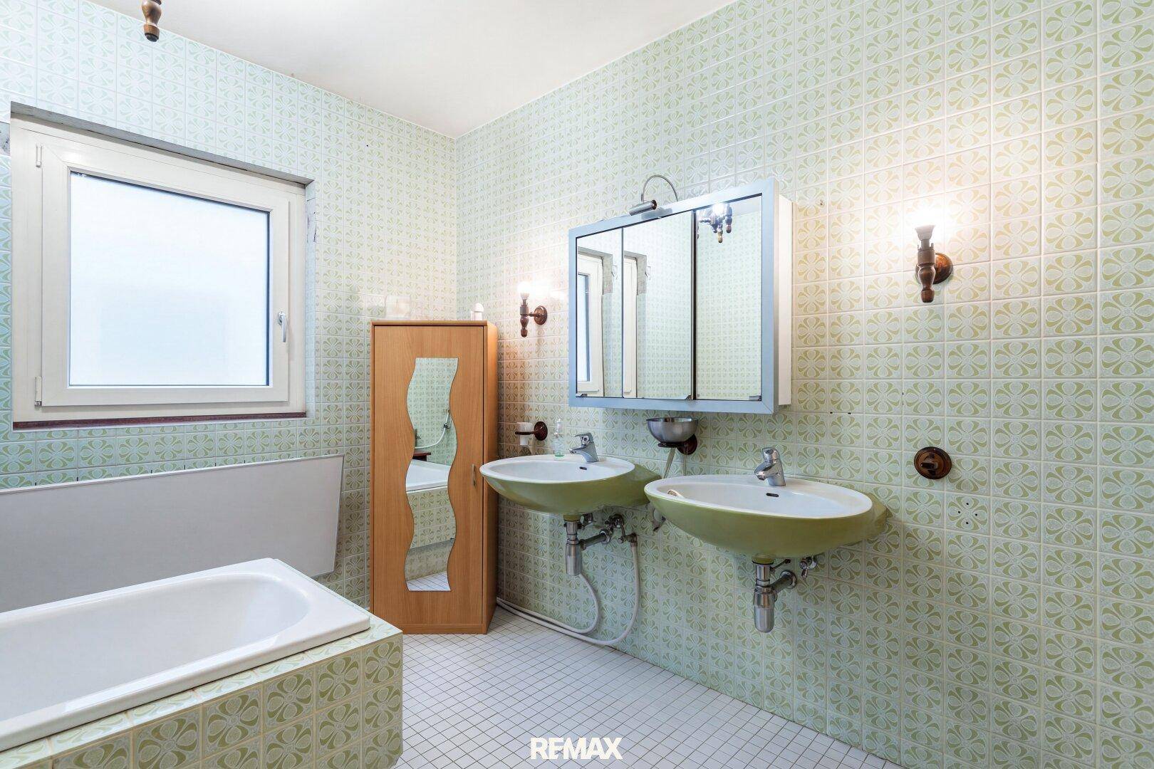 Badezimmer im 80er-Jahre-Stil mit zwei Waschbecken, Badewanne und gemusterten grünen Fliesen.