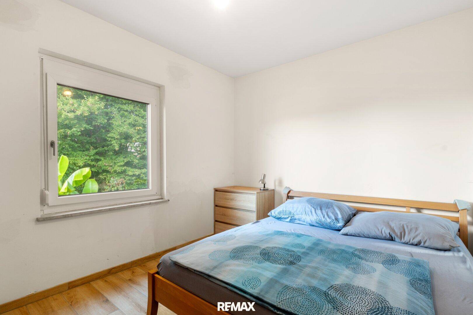 Helles Schlafzimmer mit Holzbett, Nachttisch und Fenster mit Blick auf üppiges Grün.