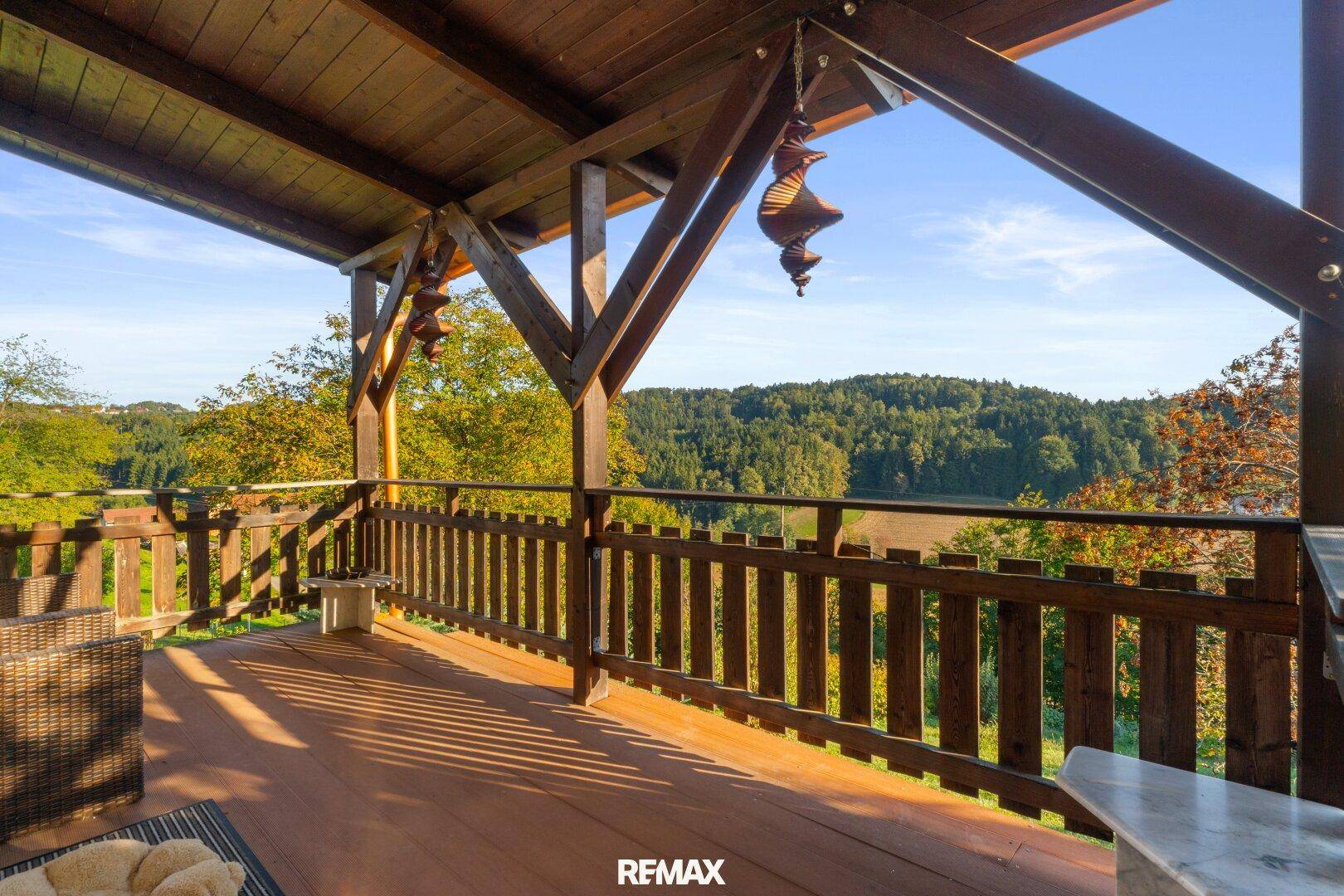 Große überdachte Terrasse mit Holzboden und Geländer, bietet einen weiten Blick ins Grüne.