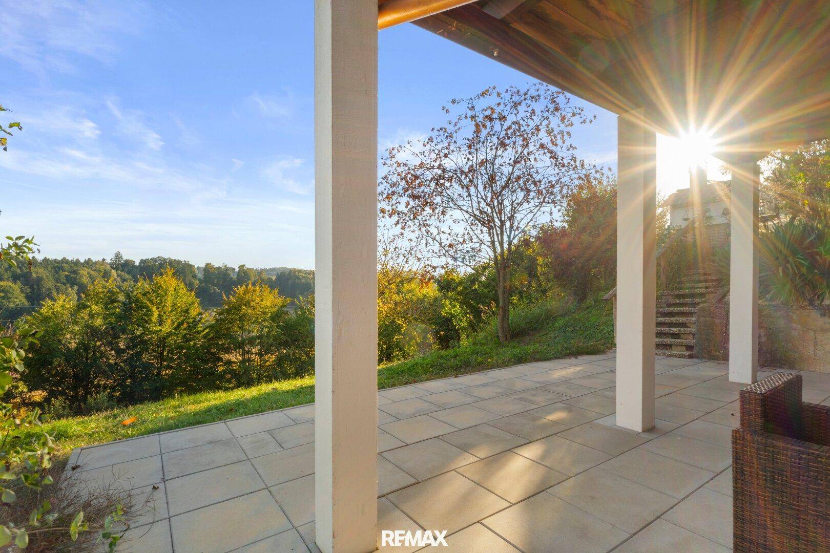 Überdachte Terrasse mit Steinplattenboden und Säulen, bietet einen sonnigen Blick ins Grüne.