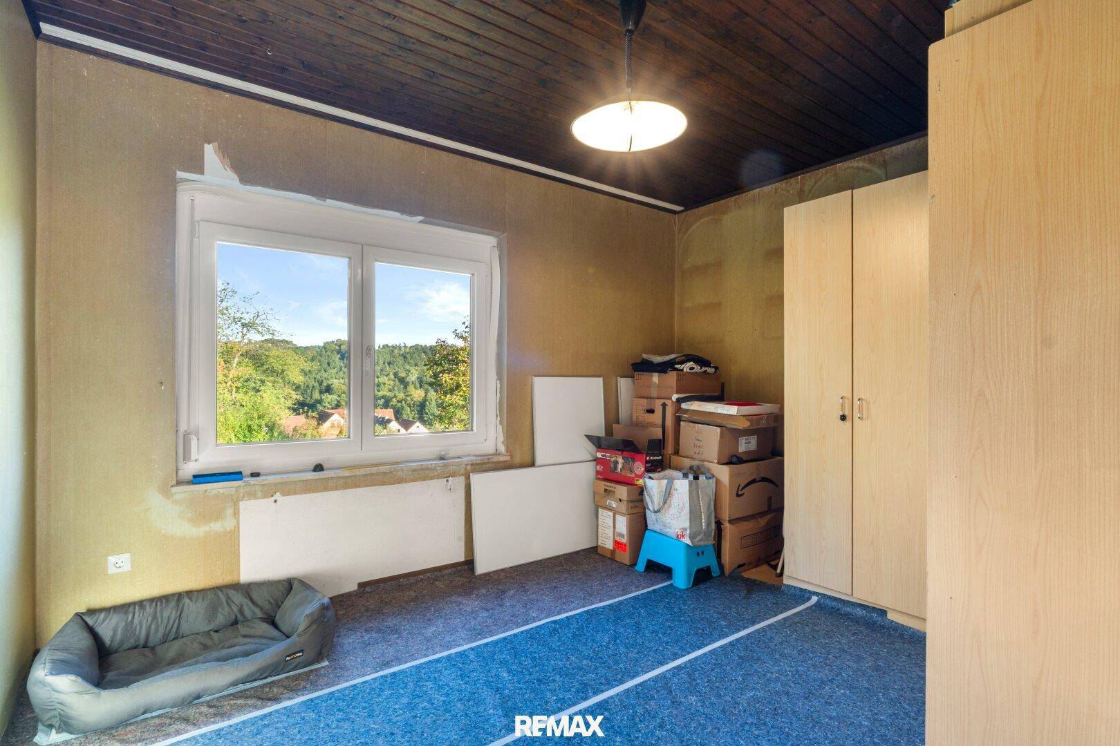 Zimmer mit Teppichboden, Holzdecke und Fenster, das Renovierungsbedarf aufweist.