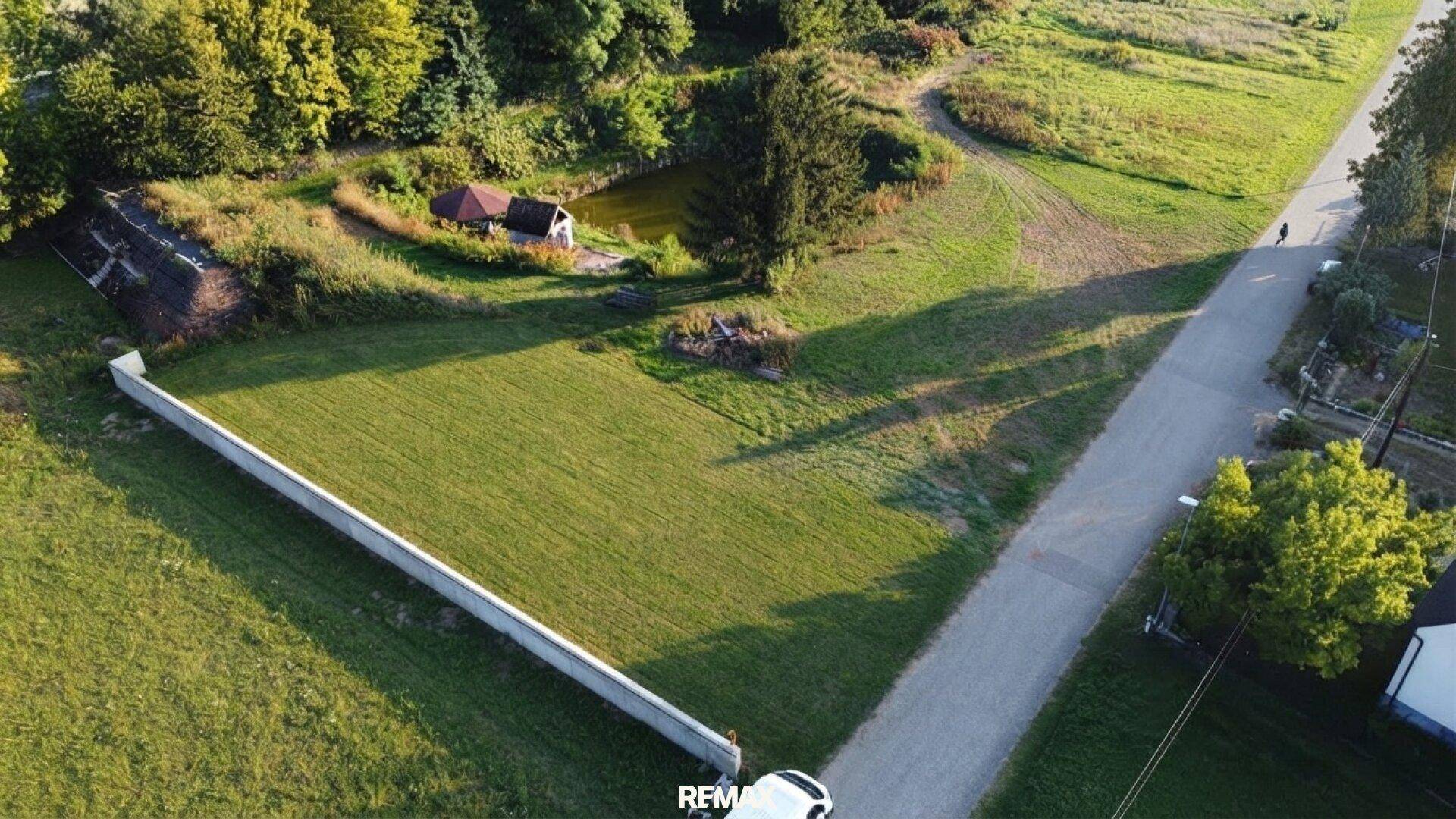 Luftaufnahme eines grünen Grundstücks mit einer langen weißen Mauer, angrenzend an eine Straße und einen Teich.