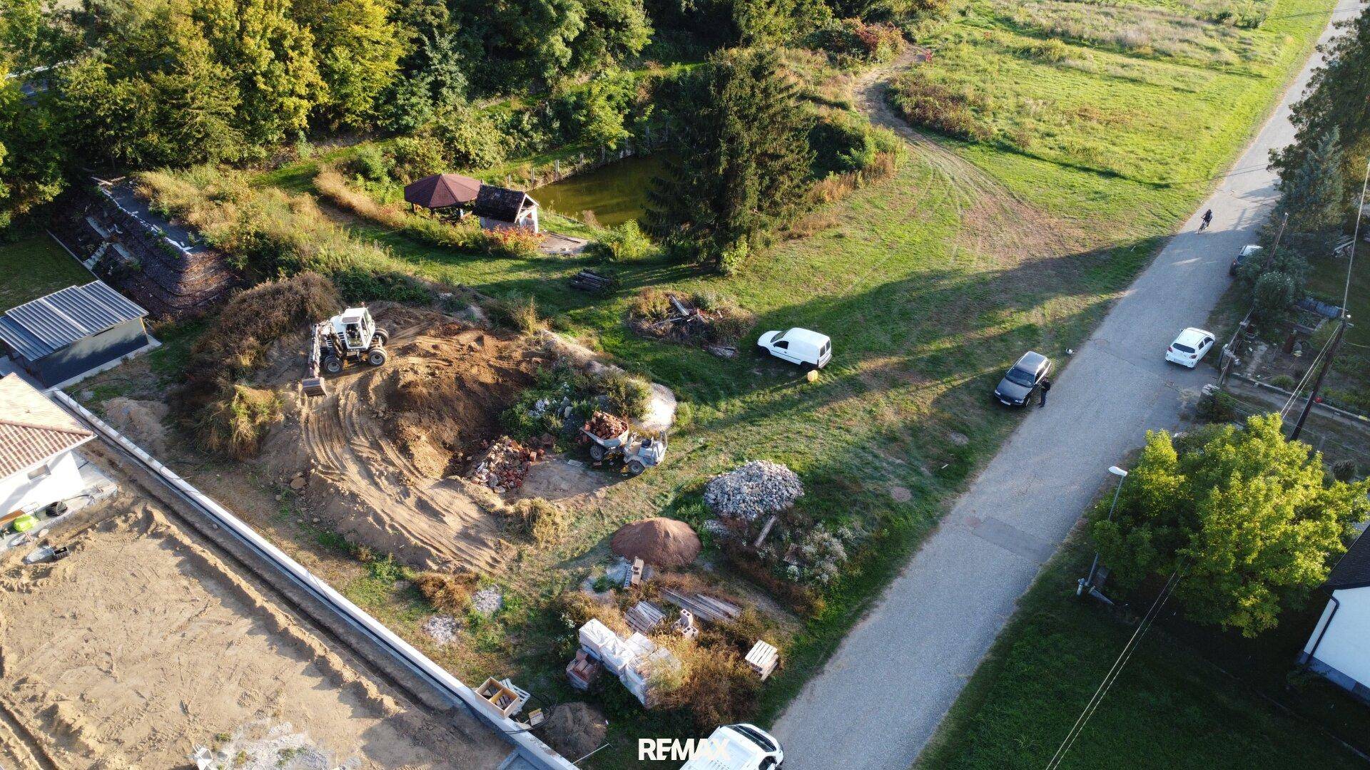 Luftaufnahme eines Grundstücks mit Bauarbeiten, Bagger und Erdhaufen, umgeben von grüner Landschaft.