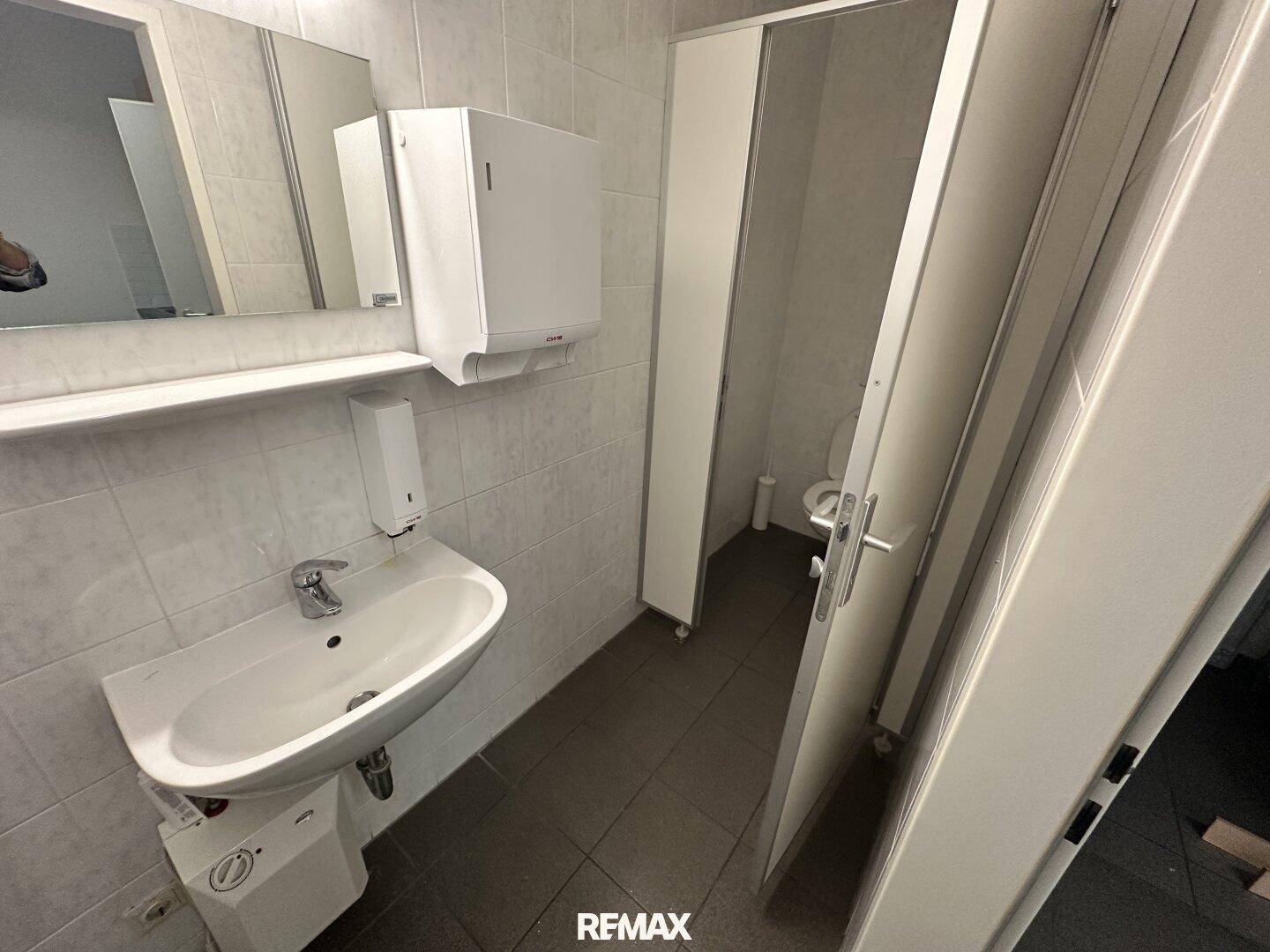 Einfacher Toilettenraum mit Waschbecken, Spiegel und gefliesten Wänden in hellem Design.