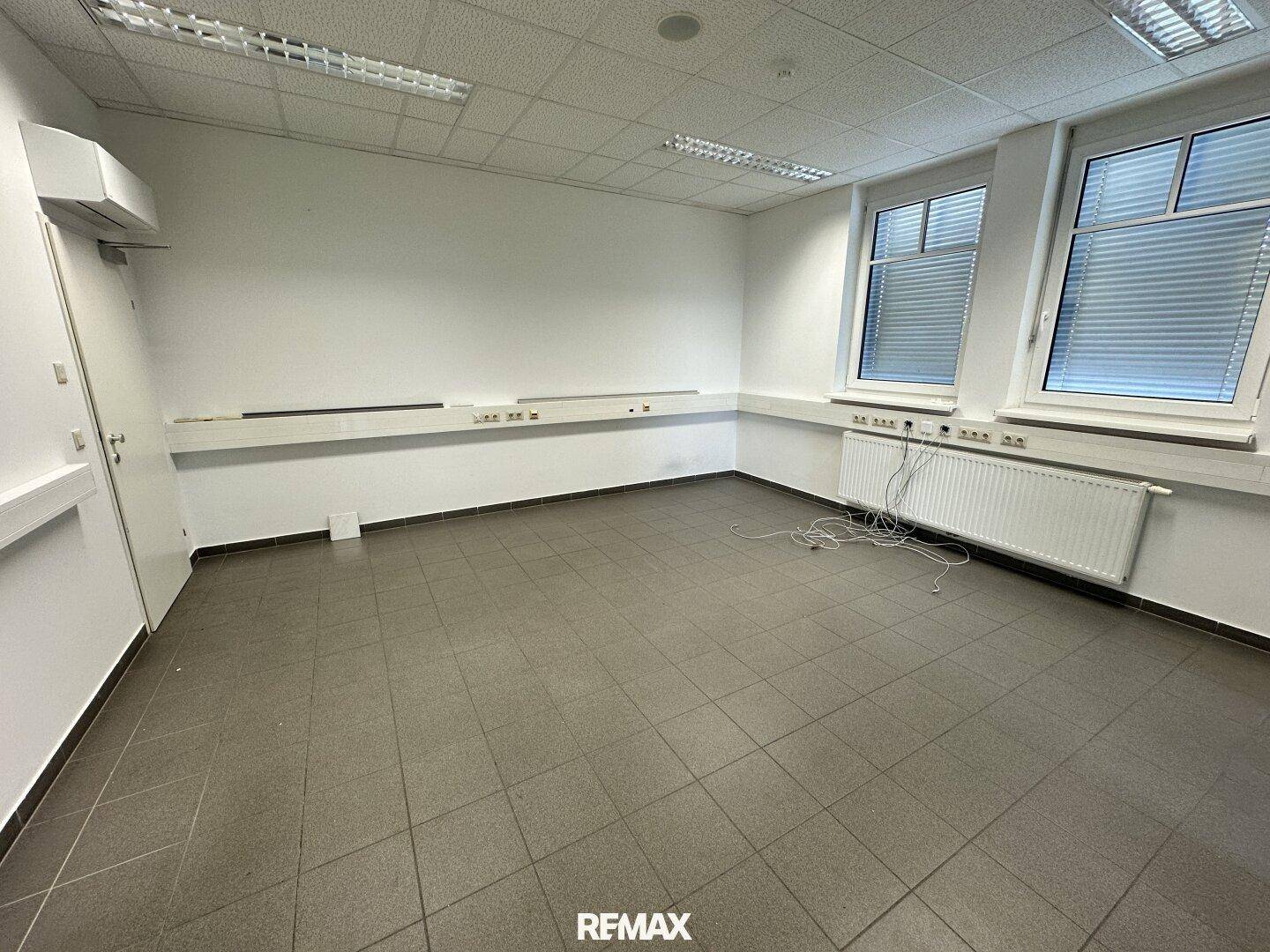 Geräumiges Büro mit zwei Fenstern, Heizkörpern und Kabeln am Boden, bereit zur Nutzung.