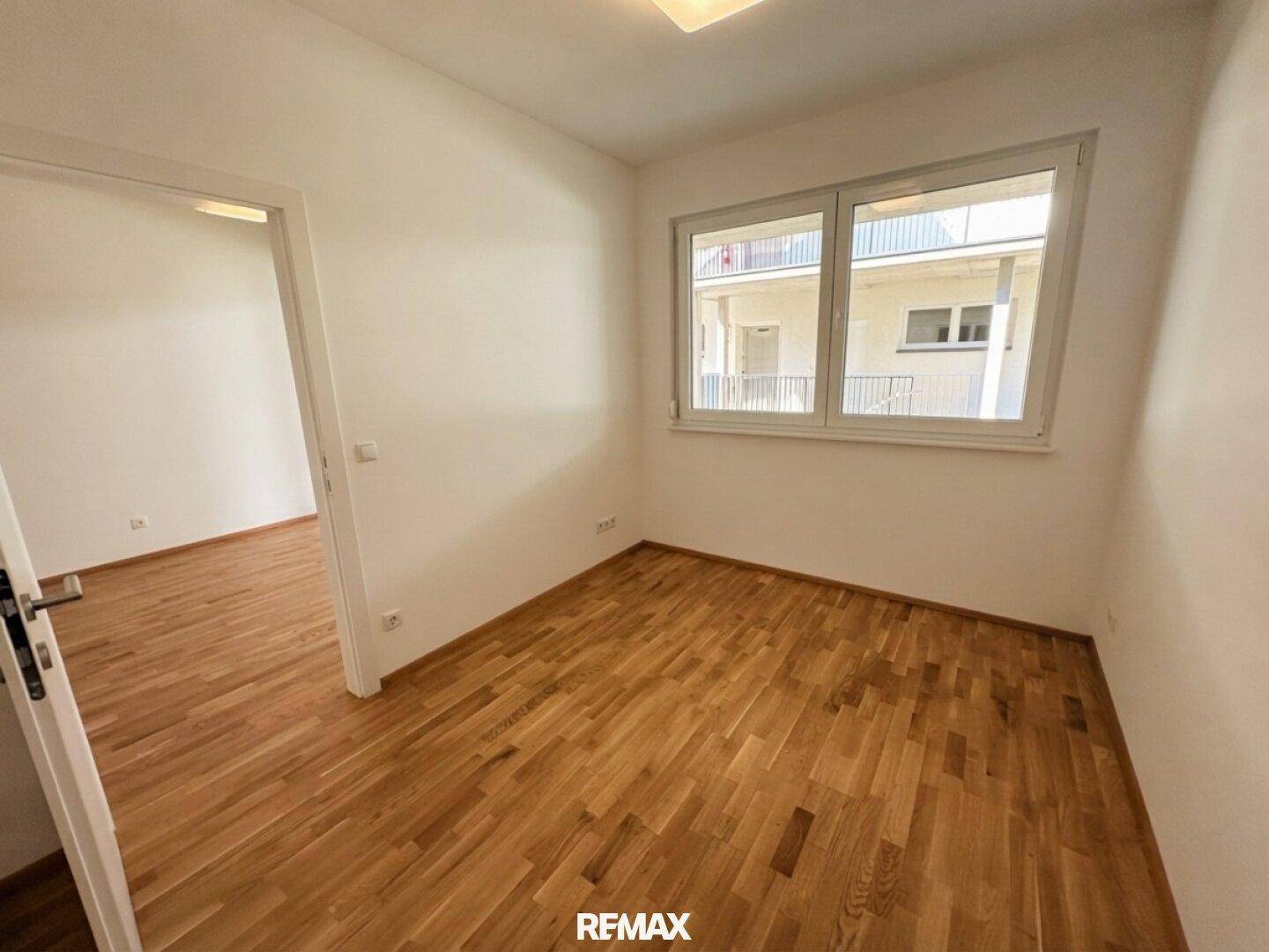 Helles Zimmer mit Parkettboden, Fenster und offener Tür zu einem angrenzenden Bereich.