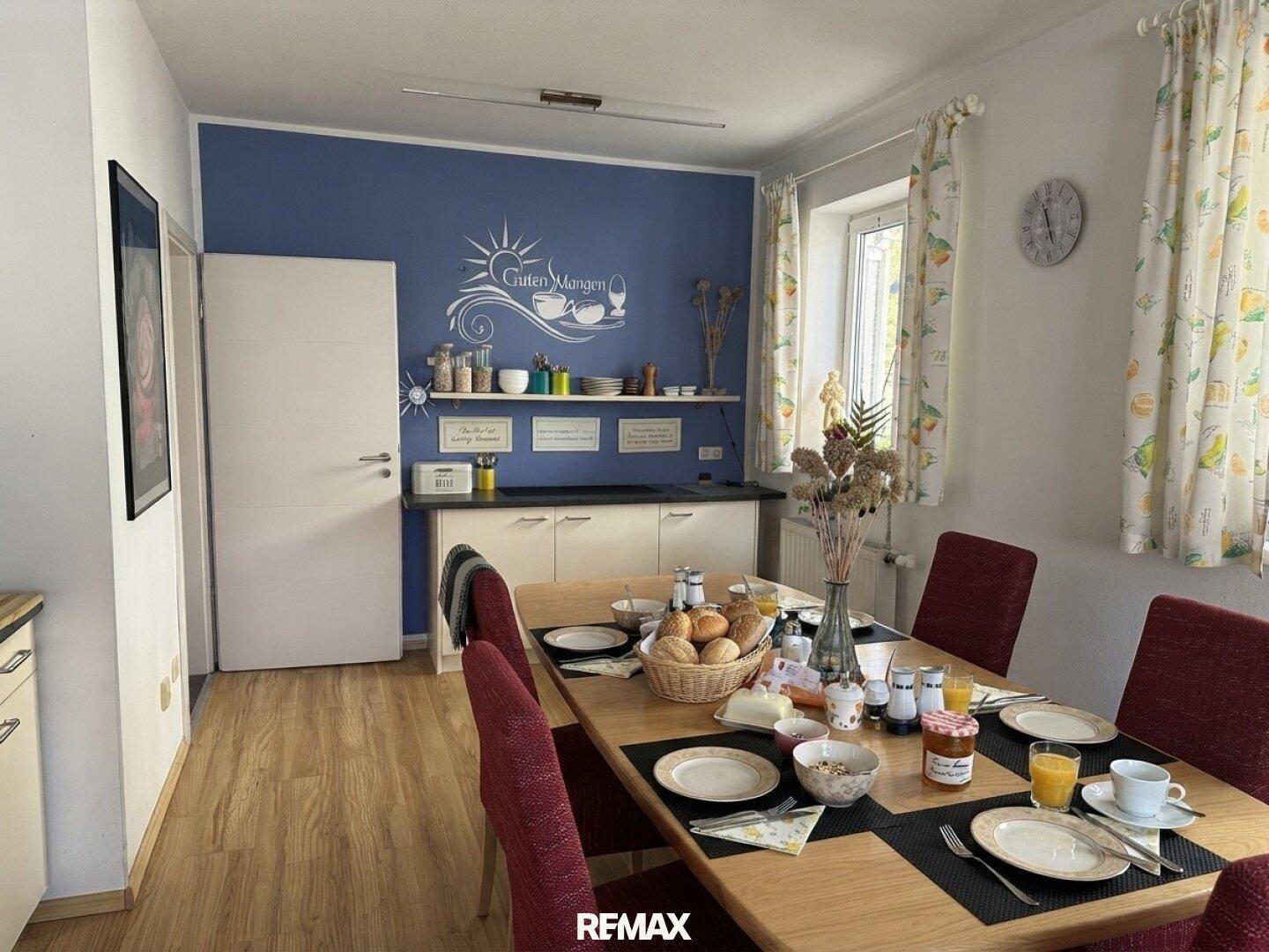 Heller Essbereich mit Holztisch, blauer Akzentwand und kleiner Küchenzeile.